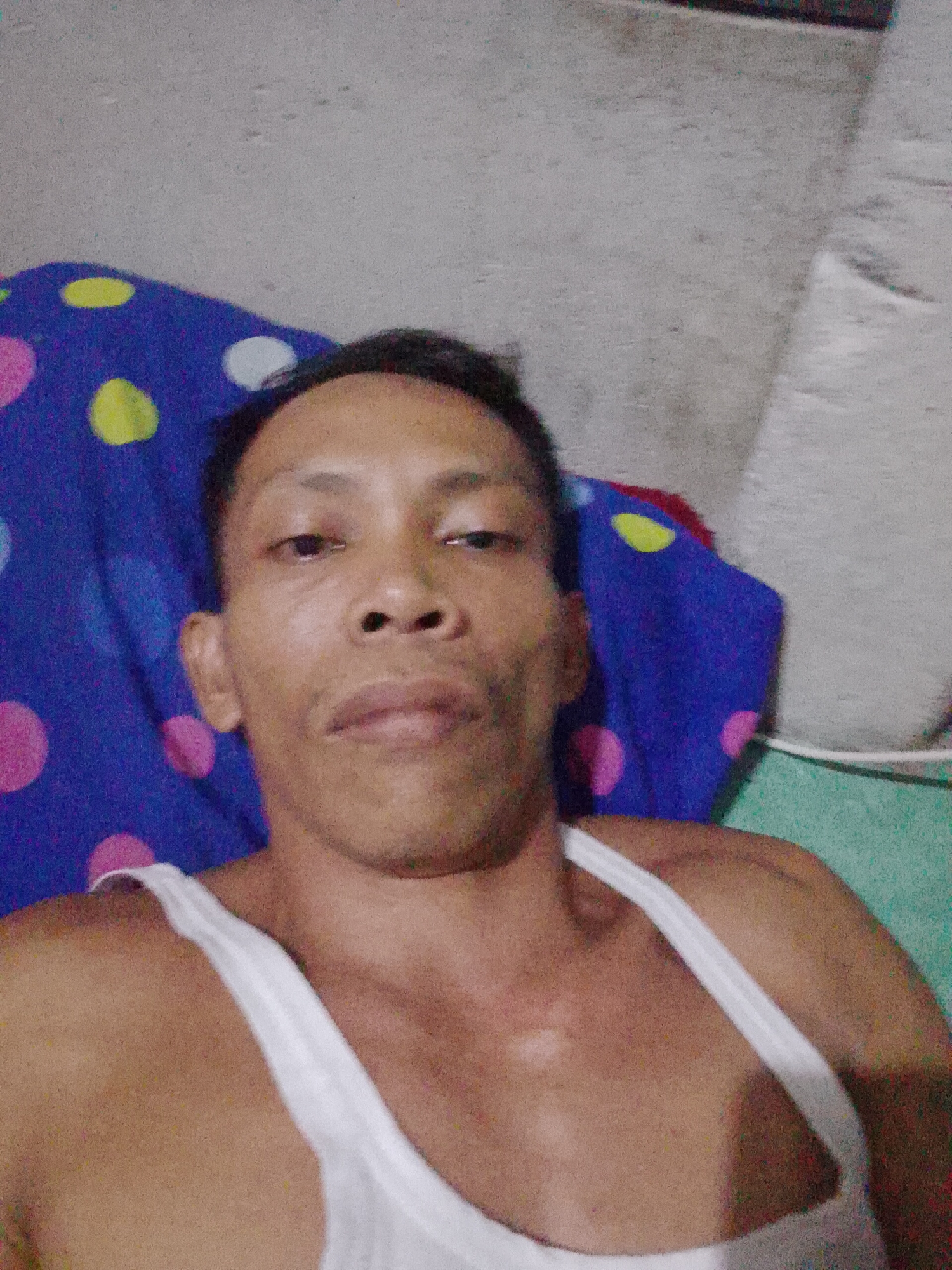A medium-shot selfie photograph taken in Bukit Ketok, Indonesia, shows a dark-skinned man wearing a white tank top, lying on his back. Behind him is a blue blanket with pink, yellow, and white polka dots. A white pillow sits to the man's right side against a light gray wall. The lighting is poor, resulting in a grainy texture. The man is looking directly at the camera with a neutral expression.