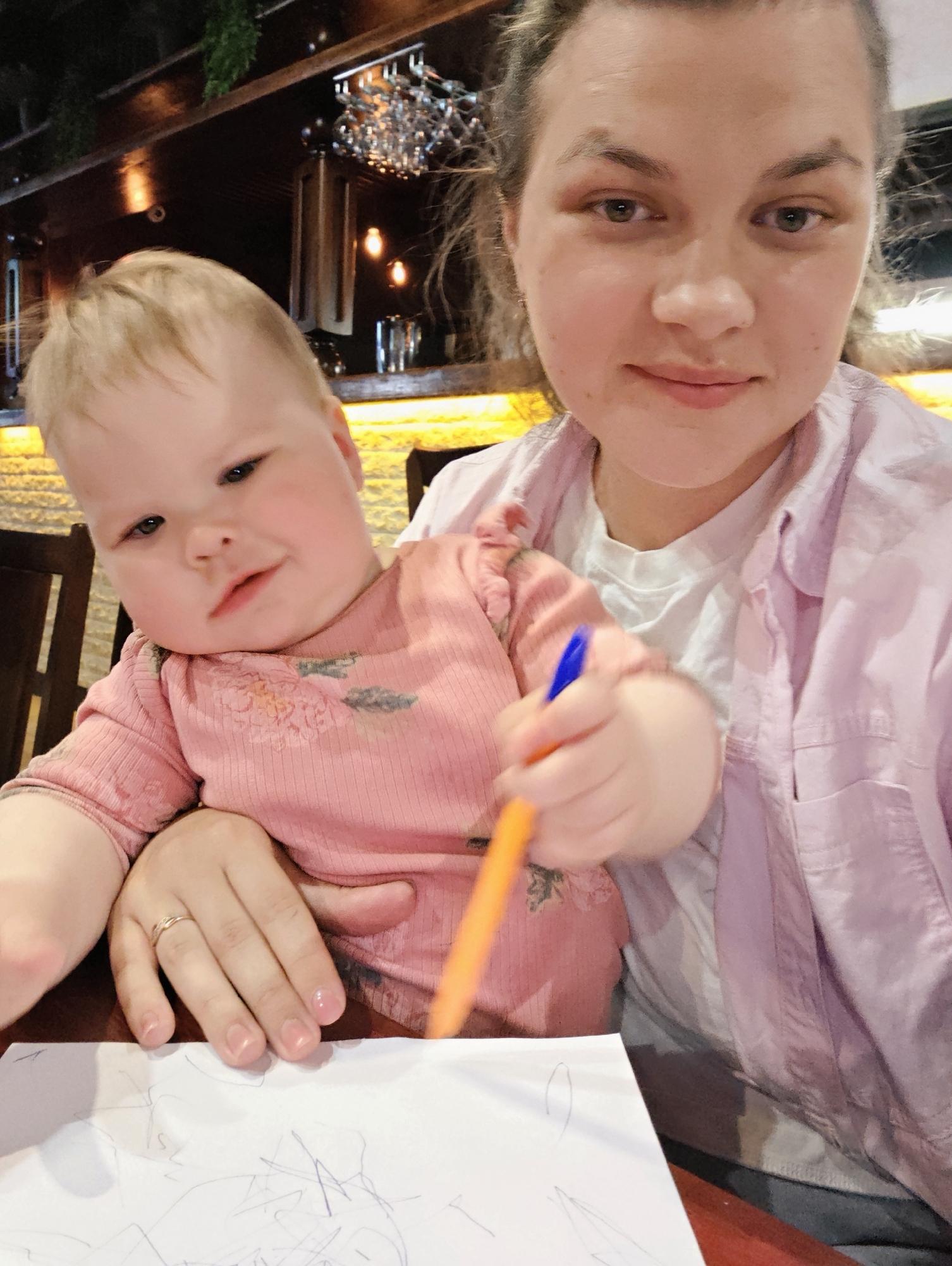 A medium, indoor shot shows a fair-skinned woman holding a fair-skinned child as the child scribbles on a sheet of paper with a pen. The woman is in the upper right of the shot, facing forward, with her hair pulled back. She wears a white t-shirt beneath a pale purple button-down shirt. The child is in the center left of the shot. They are wearing a pink, long-sleeved shirt with floral details. The child is holding an orange pen with a blue tip, and they are using it to make marks on a white sheet of paper. The child's left hand, featuring a gold ring on the ring finger, rests on the paper. The scene appears to be in a restaurant; the background shows dark wood furniture and some decorative lighting. The location may be in the city of Chaikovsky, Russia.