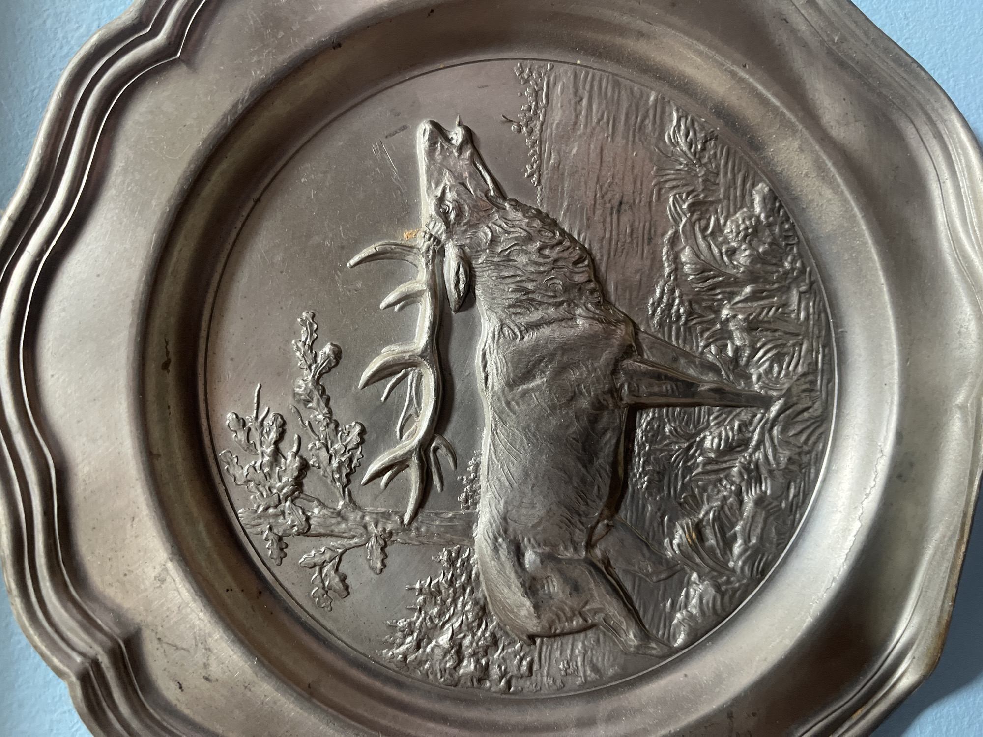 This image shows a close-up of a decorative metal plate with a bas-relief scene of a stag. The stag is depicted in profile, facing right, with its mouth open as if roaring or calling. It has large antlers and is standing in a wooded area with a tree behind it and water or a field in front. The plate has a scalloped edge. The object appears to be made of pewter or a similar metal. The background is a light blue surface. No specific location in Schwäbisch Hall, Germany, can be identified from this image.
