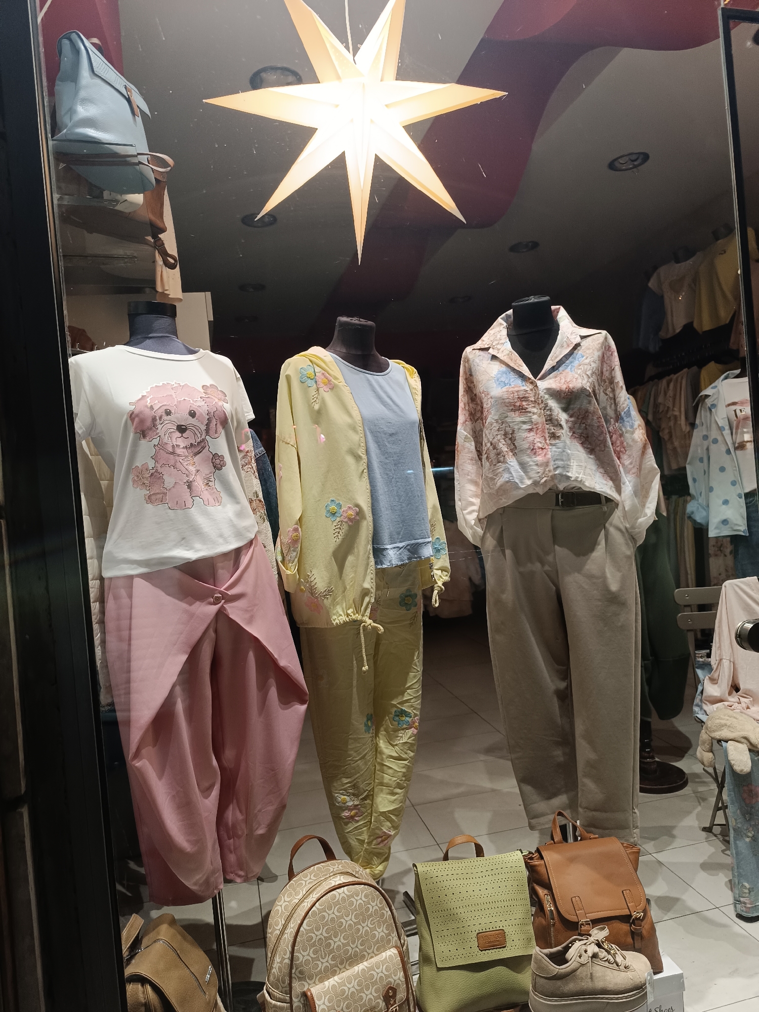 The image displays a brightly lit clothing store window showcasing three women's outfits on mannequins, likely captured at night or indoors due to prominent reflections on the glass. The reflections include a red and black wavy ceiling design, recessed lights, and some dust.
On the far left, a mannequin wears a white short-sleeved top featuring a large, textured pink fluffy dog graphic adorned with small flowers. This is paired with light pink, wide-legged draped pants that have a buttoned detail on the front.
The central mannequin is dressed in a light blue top, layered with a pastel yellow long-sleeved jacket and matching yellow pants. Both the jacket and pants are embellished with delicate floral embroidery in shades of blue and pink.
To the right, the third mannequin wears a loose-fitting, long-sleeved collared shirt with a subtle floral and abstract pattern in light blue, pink, and beige tones. This shirt is paired with high-waisted, beige pleated trousers secured with a black belt.
Below the mannequins, an array of accessories is displayed. These include a light beige backpack with a repeated "C" pattern, a lime green perforated backpack with a label possibly reading "ARTGE", and a tan leather-like backpack. A pair of beige platform sneakers rests on a shoebox, partially revealing the text "e Shoes". Higher up on the left, a light blue shoulder bag and a brown messenger bag are also visible. A large, prominent star-shaped light fixture hangs from the ceiling, illuminating the display. The background inside the store shows blurred clothing racks. The setting is a retail display window in Pleven, Bulgaria.