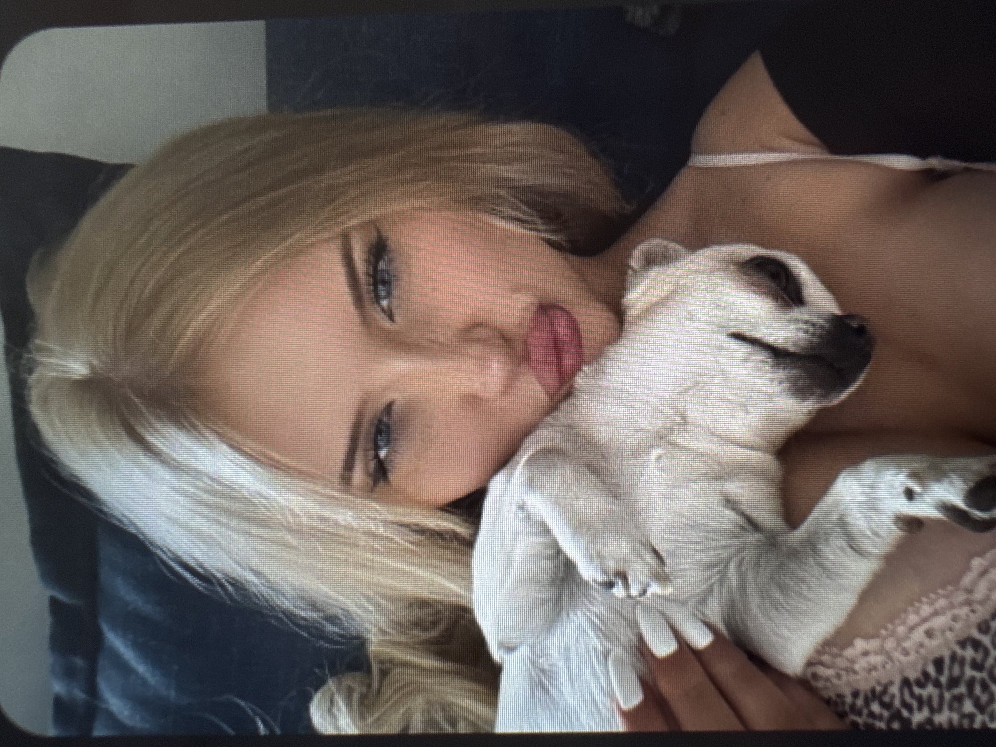 The image captures a blonde woman lying down, holding a small, light-colored dog. Her head is tilted to the side, and she looks directly at the viewer with blue eyes, sporting a pouty expression with slightly parted, reddish lips. Her long blonde hair is swept back, and she appears to be wearing a light-colored, possibly lace-trimmed top or bra. Her hands, visible near the dog, show long, light-colored fingernails.
The small dog, which has light, possibly white or cream-colored fur, is nestled against the woman's chest and neck area. Its head is turned away from the camera, and its eyes are not distinctly visible, suggesting it is resting comfortably. Part of a leopard print fabric is visible in the lower right corner, underneath the dog and the woman's arm.
The background is dark and appears to be bedding or a pillow. The scene evokes a casual, intimate moment between the woman and her pet. The image itself has a noticeable grainy or pixelated quality, with a visible grid-like pattern across the entire frame, indicating it may be a photograph taken of a screen. The corners of the image are slightly rounded. There is no visible text.