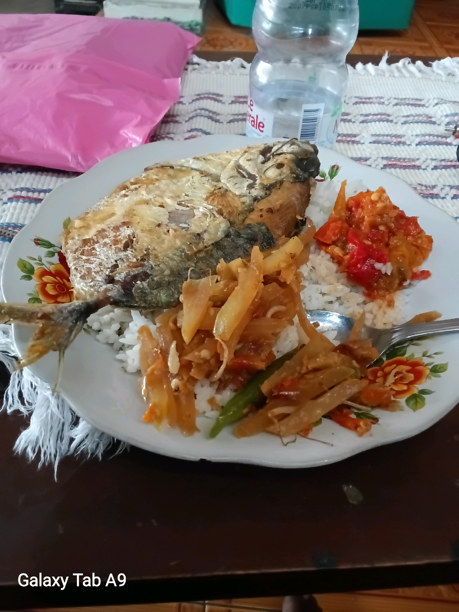 A close-up, overhead shot shows a plate of food on a patterned placemat. The plate is white with floral designs and contains a portion of fried fish, white rice, a spicy red chili sauce, and a side dish of sliced vegetables. A fork rests on the plate. In the background, a clear plastic bottle of water is visible. To the left of the plate is a bright pink plastic bag, and the table surface is dark brown wood. The overall setting suggests a casual meal. The image also displays the text "Galaxy Tab A9" in the bottom left corner. This scene is likely located in a household or local eatery in Trenggalek, Indonesia, given the commonality of such dishes and the prevalence of the Samsung Galaxy Tab A9 device for casual photography in the region.