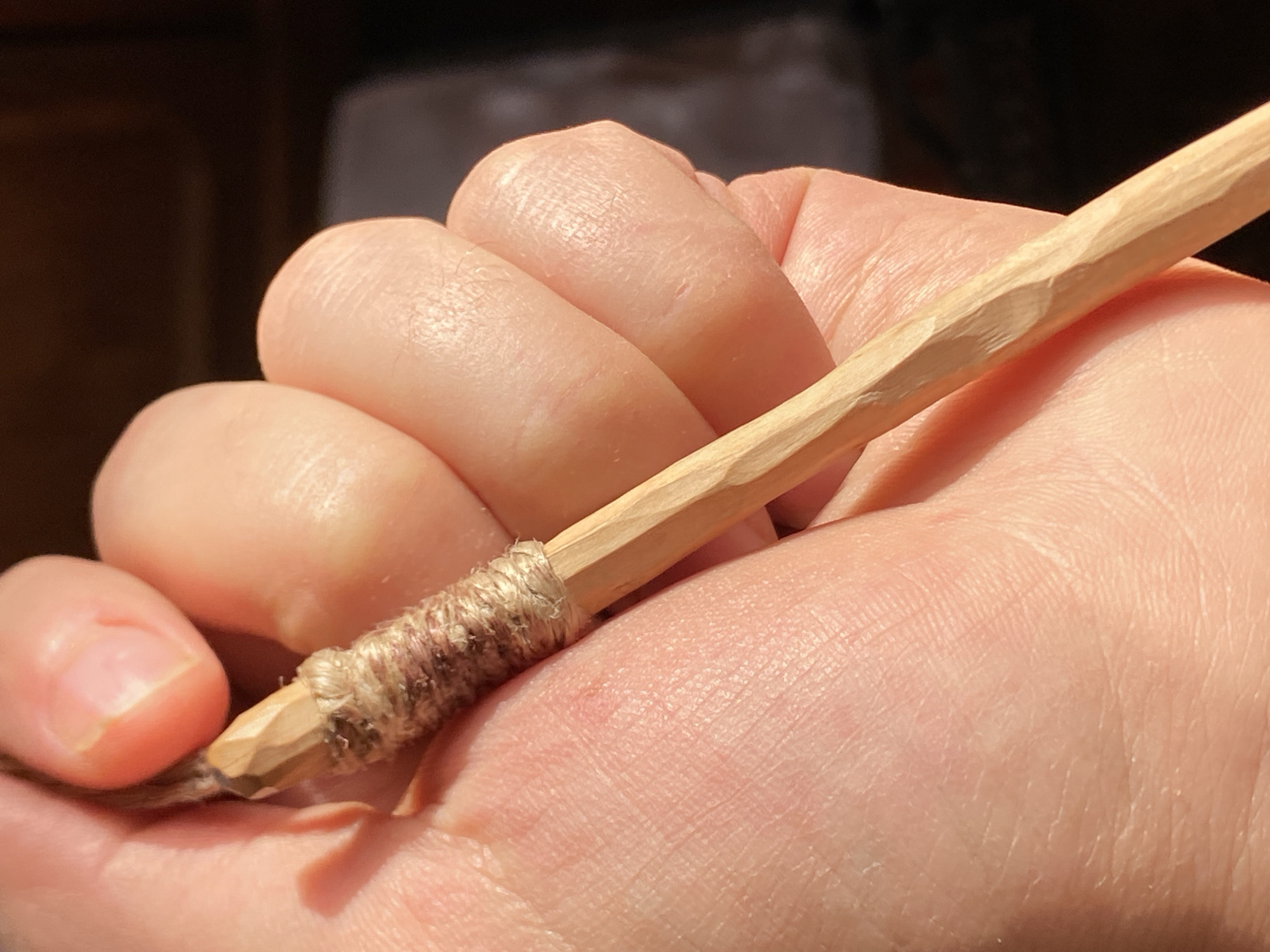 A close-up, low-angle shot shows a hand holding a sharpened wooden stick. The stick is wrapped with twine around its top section, just below the sharpened tip. The hand, with its light skin and visible creases, cradles the stick. The background is blurred, suggesting a shallow depth of field, with darker areas hinting at shadows and possibly a dimly lit indoor environment. The lighting casts a warm glow on the skin and wood. The scene does not contain any identifiable landmarks or elements that would indicate a specific location in Belgrade, Serbia.