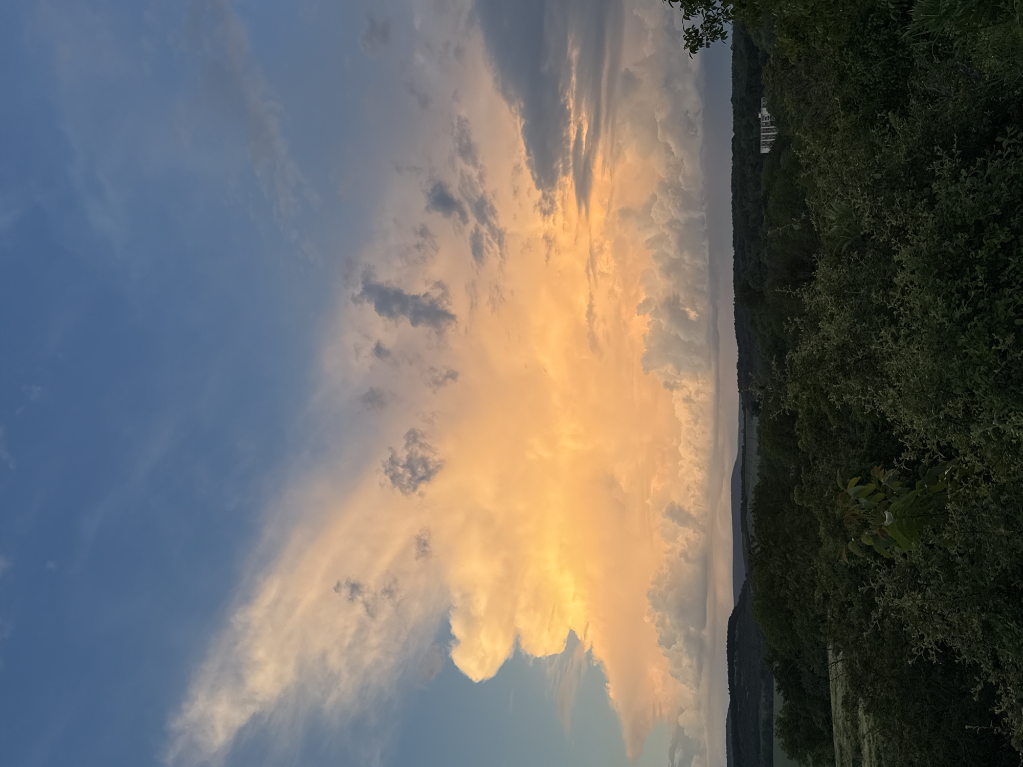 The image shows a sky with a large, bright orange and white cloud formation. The sky transitions from a deep blue on the left to lighter shades around the cloud. To the right of the cloud is a dark green band of trees, possibly a forest. Beyond the trees, the landscape is indistinct, but may include hills or valleys.
Based on the location, this image is likely taken in a rural setting near Strazhitsa, Bulgaria, with the prominent feature being the dramatic sunset or sunrise cloud formation. Buildings can be seen in the distance, behind the trees.