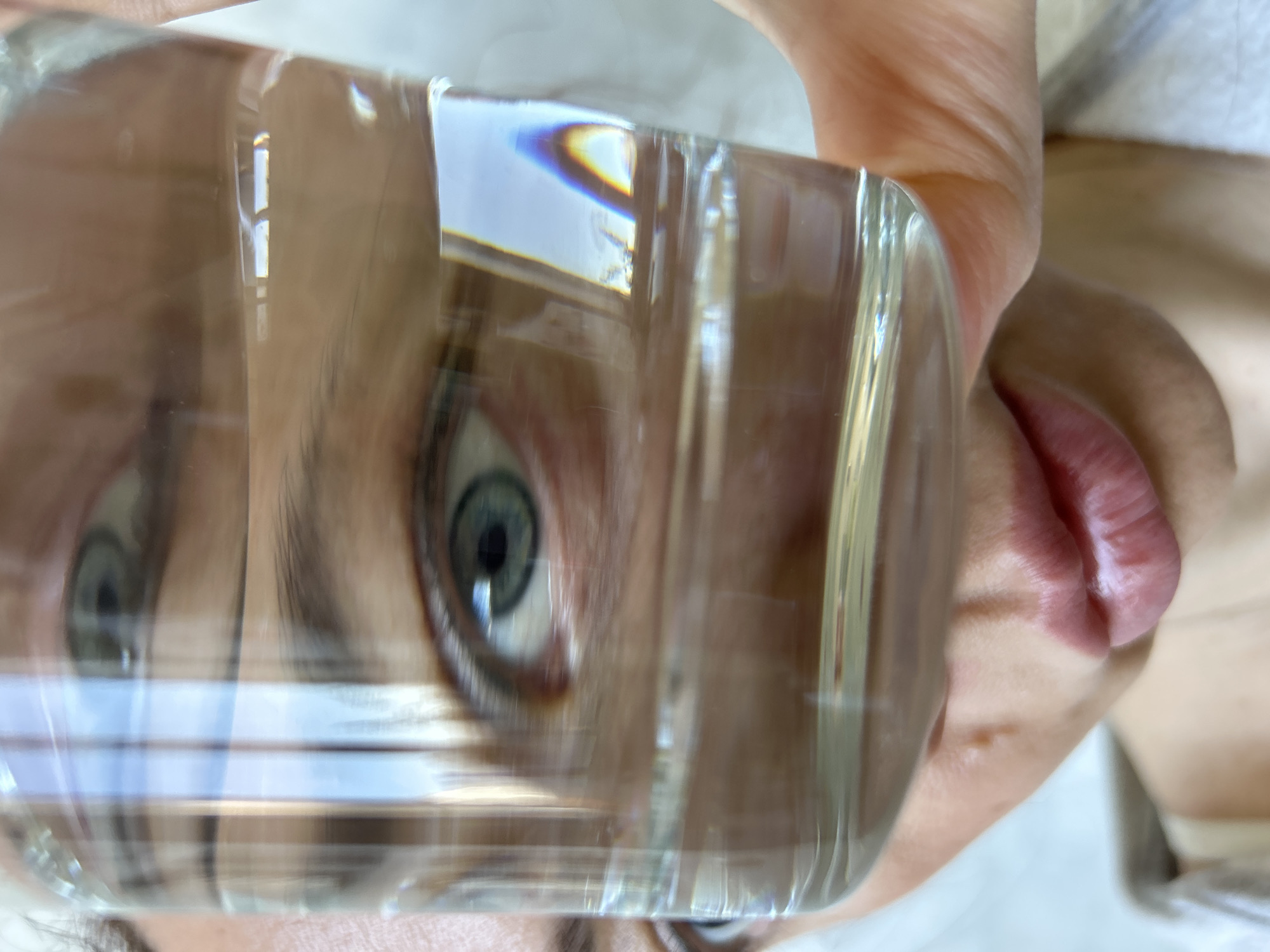 The image shows a person's face distorted by a glass object filled with liquid, likely water. The person has light skin, blue eyes, and pink lips. Their face is partially visible through the glass, with the eye and mouth being the most distinct features. The glass object is held close to the face, creating a distorted and magnified view. The background is blurry and indistinct. There is no information that indicates the location is the city of Chaikovsky, Russia.