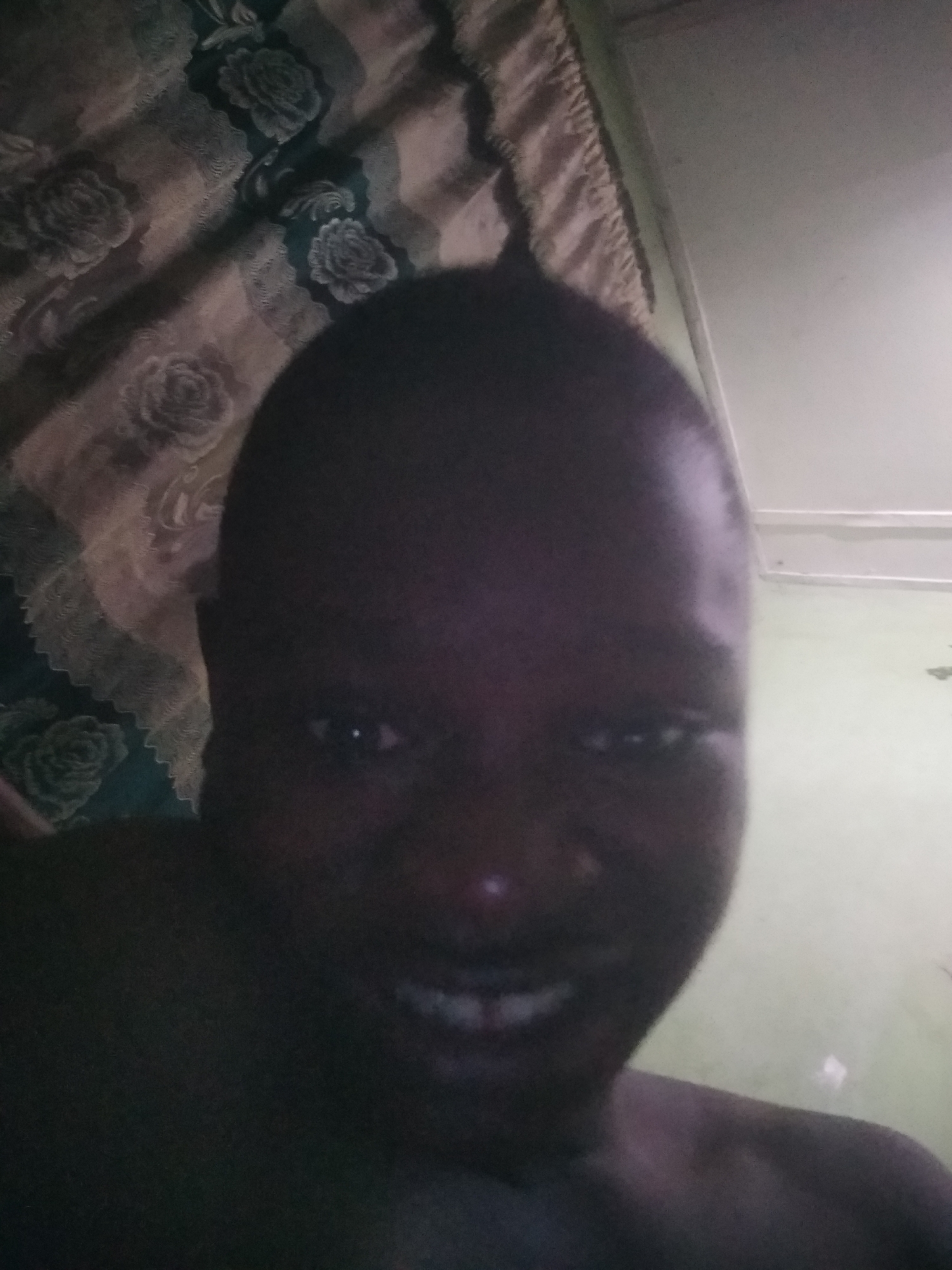 A dark, close-up selfie captures a person with dark skin, likely male, from the chest up. The person is positioned centrally, looking directly at the camera with a wide smile that reveals their teeth. Their head is mostly visible, with the top of their head appearing bald or with very short hair.
The setting appears to be indoors in Shangisha, Nigeria, characterized by low light conditions. To the left and upper-left of the person, a patterned fabric, possibly a curtain or bedsheet, is visible. It features a floral design with dark rose motifs against a lighter, possibly faded or discolored, background. To the right and upper-right, a plain light-colored surface, likely a wall or ceiling, is visible. The overall image is underexposed, making details somewhat obscured, but reflections are discernible in the person's eyes. The person's expression conveys amusement or happiness. No visible text is present in the image.