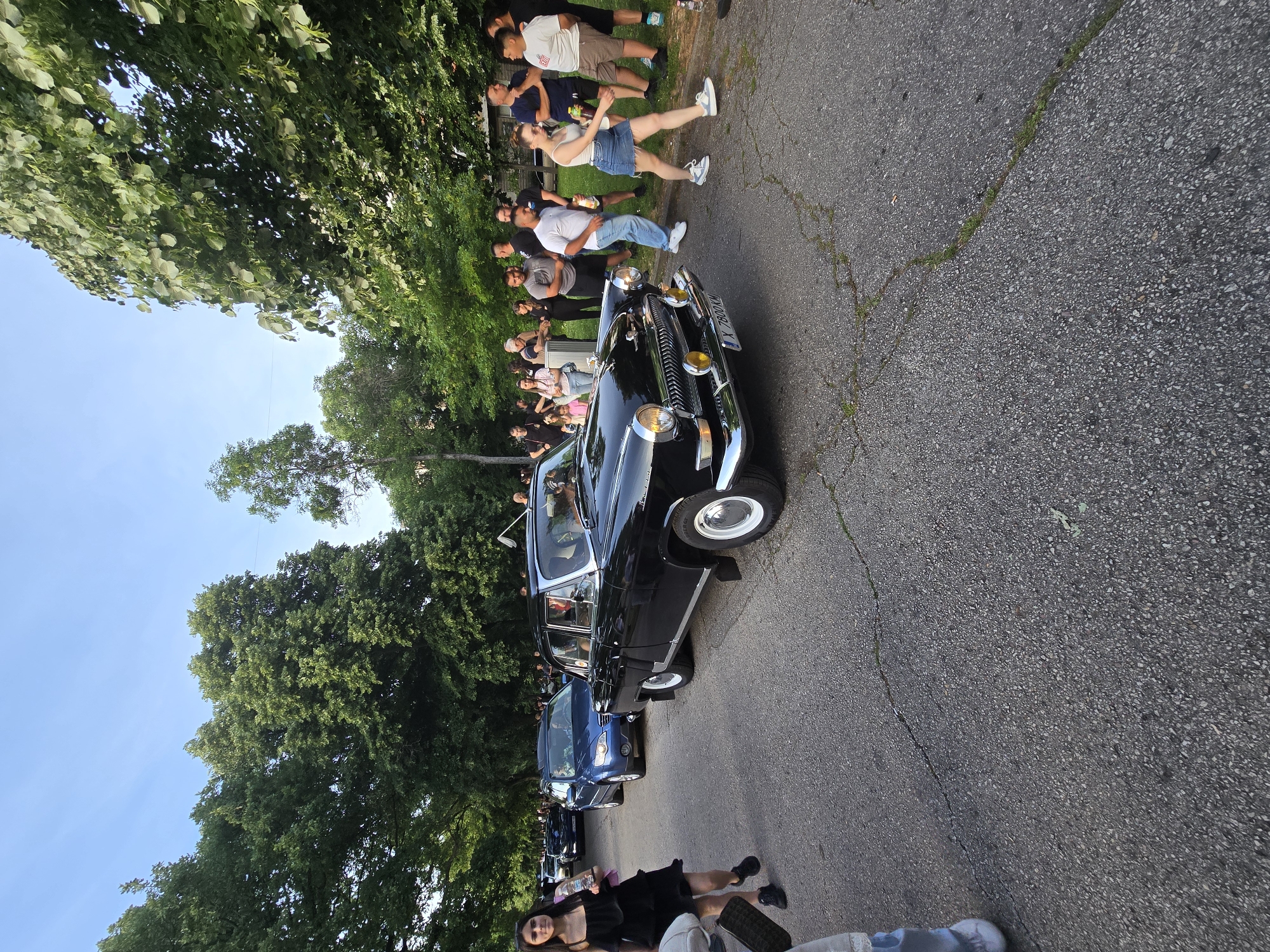 A black vintage car is driving down a cracked asphalt road lined with trees, while a crowd of people stands on the side of the road watching it pass. There are other cars parked behind the vintage car. The location appears to be a residential street.