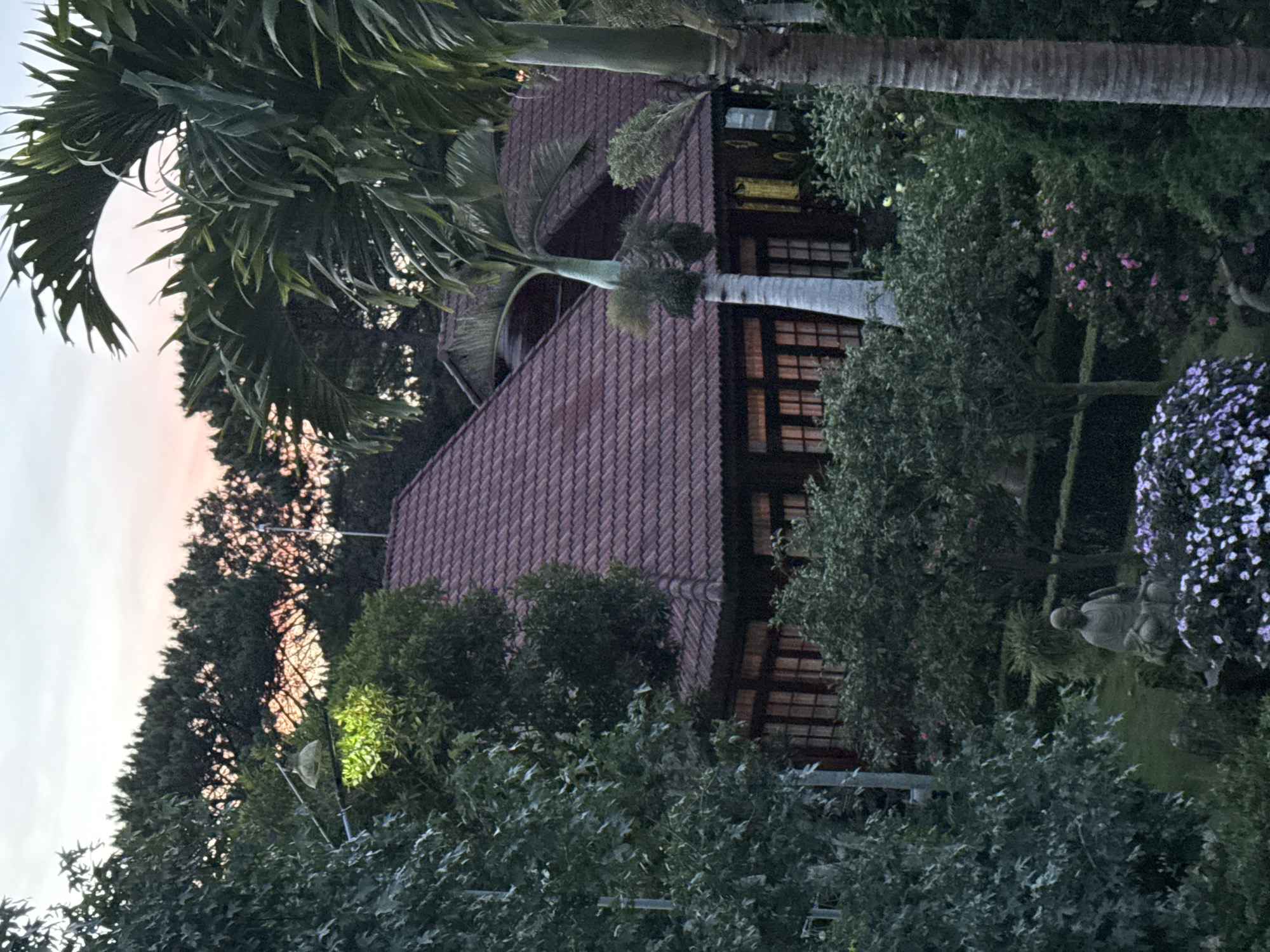 This image depicts a twilight scene at a villa in Da Lat, Vietnam. The villa features a red tiled roof and dark wooden exterior, with several windows illuminated from within. Lush greenery surrounds the property, including palm trees and manicured bushes. A statue of a seated figure is visible in the foreground, surrounded by flowering plants. The sky displays soft hues of pink and orange, indicative of sunset or sunrise.