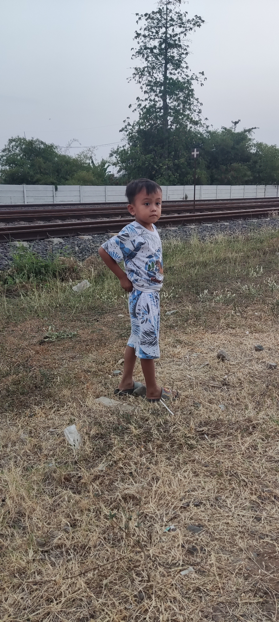 A young boy stands facing the right of the frame with his right hand on his hip. He is wearing a white t-shirt with a blue and white leaf pattern and blue and white leaf-patterned shorts. He is wearing dark-colored flip-flops. The boy is standing on dry, yellowed grass and a few rocks. Behind him, there are two sets of railroad tracks, with gravel between them. A low, white concrete barrier runs along the edge of the tracks. Further in the background, there are green trees and a tall, slender tree with sparse leaves. A white sign with a red cross on it is visible among the trees. The sky is overcast and gray. The scene appears to be located outdoors, possibly near a rural area or the outskirts of a town, given the railroad tracks and natural vegetation. The lighting suggests it is either early morning or late afternoon. The location is identified as Pakusamben, Indonesia.