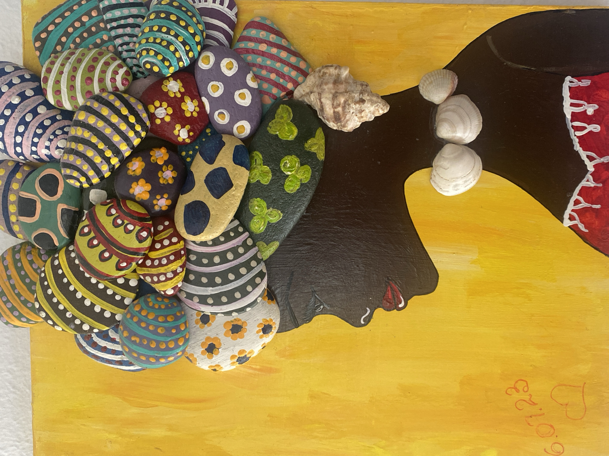 The image shows a painting of a woman’s head in profile, created using rocks and shells. The woman's face and neck are painted black, with a red lip and white details defining her eye. Her hair is represented by a collection of painted rocks with various colors and patterns, clustered together. There are also seashells added to the hair. The background is a solid yellow color. In the bottom right corner, there is a signature that reads "6.01.23" with a heart shape. The painting is likely located in Çekmeköy, Turkey, based on the search query including this location. There are no other people depicted or interactions described.