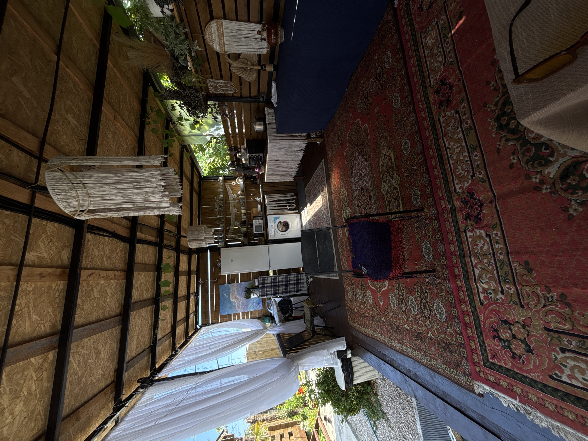This image depicts a covered outdoor seating area with a rustic, bohemian aesthetic. The ceiling and walls are constructed from natural wood, with exposed beams and wooden paneling, suggesting an open-air or semi-open structure.
The scene is furnished with a variety of items, creating a lived-in and eclectic feel. A large, ornate red Persian rug covers a significant portion of the floor, its intricate patterns adding a touch of traditional grandeur. Overlaid on this is a dark blue runner. Seating is provided by a metal chair draped with a dark blue and red cushion. Nearby, a small table with a checkered cloth holds various objects.
Towards the back of the space, there appears to be a small kitchenette area, with a white refrigerator and a washing machine visible. Decorative elements include macrame hangings and various plants, some trailing from above and others placed on shelves. A painting hangs on the wall, depicting a cloudy sky. White sheer curtains are draped at the entrance, allowing natural light to filter in.
The overall impression is one of a relaxed, bohemian outdoor living space, possibly a lounge area or a creative studio. The lighting suggests it is daytime, with sunlight casting shadows and highlighting textures. The location context of Gudauta, Abkhazia, implies a potentially coastal or resort-like setting, although specific location cues are not strongly evident within the frame itself. There is no visible text in the image. The atmosphere conveyed is one of comfort, creativity, and a connection to nature.
