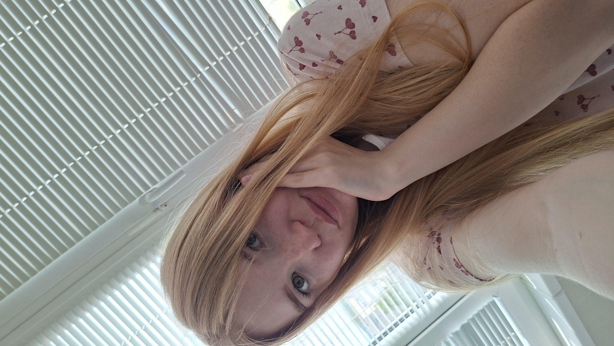 The image features a young woman with long, light reddish-blonde hair, light-colored eyes, and visible freckles on her face. She is looking directly at the camera, her expression appearing somewhat neutral or pensive. One of her hands is positioned near her face, with fingers resting on her cheek, partially covering her mouth. Her hair is spread around her, covering parts of her shoulders and arms.
She is wearing a light-colored garment, possibly a top or a dress, adorned with a repeating pattern of small, dark red heart shapes. In the background, there are white window blinds that are mostly closed, creating a distinct pattern of horizontal lines and shadows. Bright natural light filters through the blinds, suggesting it is daytime. The image is angled diagonally, indicating it might be a selfie taken from an overhead perspective while she is lying down or reclining.
The scene takes place indoors, likely in a room, with the prominent white blinds suggesting a window nearby. No specific textual elements are visible within the image. The location context provided is Chaikovsky, Russia.