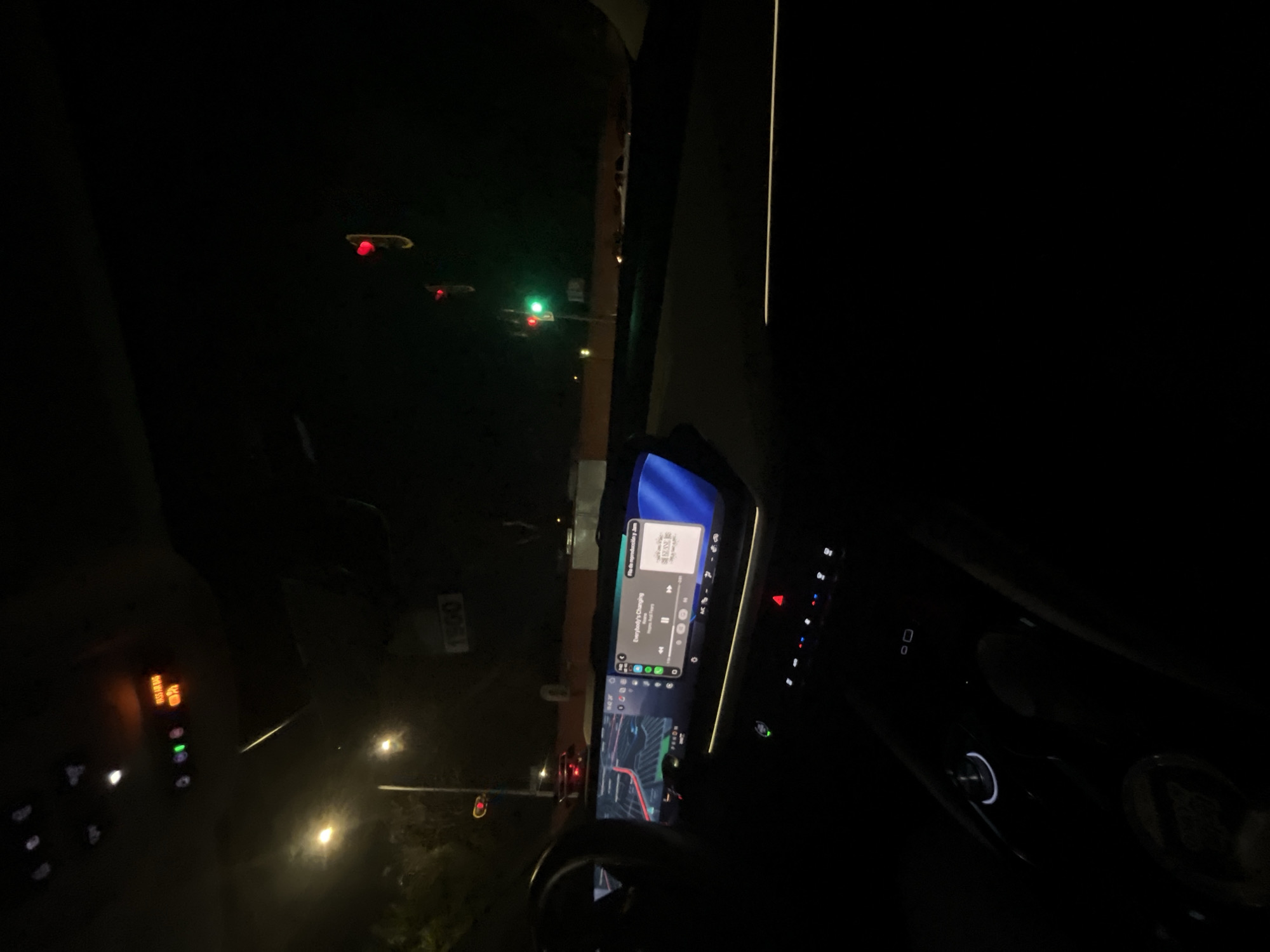 The scene depicts a nighttime view from the interior of a vehicle stopped or moving slowly on an urban street in Puebla, Mexico. The perspective is from the driver's or front passenger's seat, looking forward through the windshield.
**Vehicle Interior:**
The dashboard features a prominent, elongated dual-screen infotainment system. The left screen displays a navigation map with a route highlighted in red. The right screen shows a music player interface, indicating "Everybody's Changing" by Keane is playing, with Spanish text "Fin de reproducción y más" at the top right. Below the infotainment screen, illuminated climate controls and other buttons, including a red hazard light indicator, are visible. The center console includes a rotary control dial. The overhead console has illuminated indicators, including green lights and an orange "PASS AIRBAG OFF" warning with a passenger icon. A portion of the steering wheel is visible on the left.
**Exterior Environment:**
Outside the vehicle, the street is dark, illuminated by multiple streetlights casting yellow-white light. Several traffic lights are present: one is green directly ahead, indicating permission to proceed, while another appears red in the distance to the upper right. A dark rectangular sign with "1800" in white text is visible in the mid-ground. The background is predominantly dark, with a light-colored wall or structure discernible in the far distance. No individuals are explicitly visible.