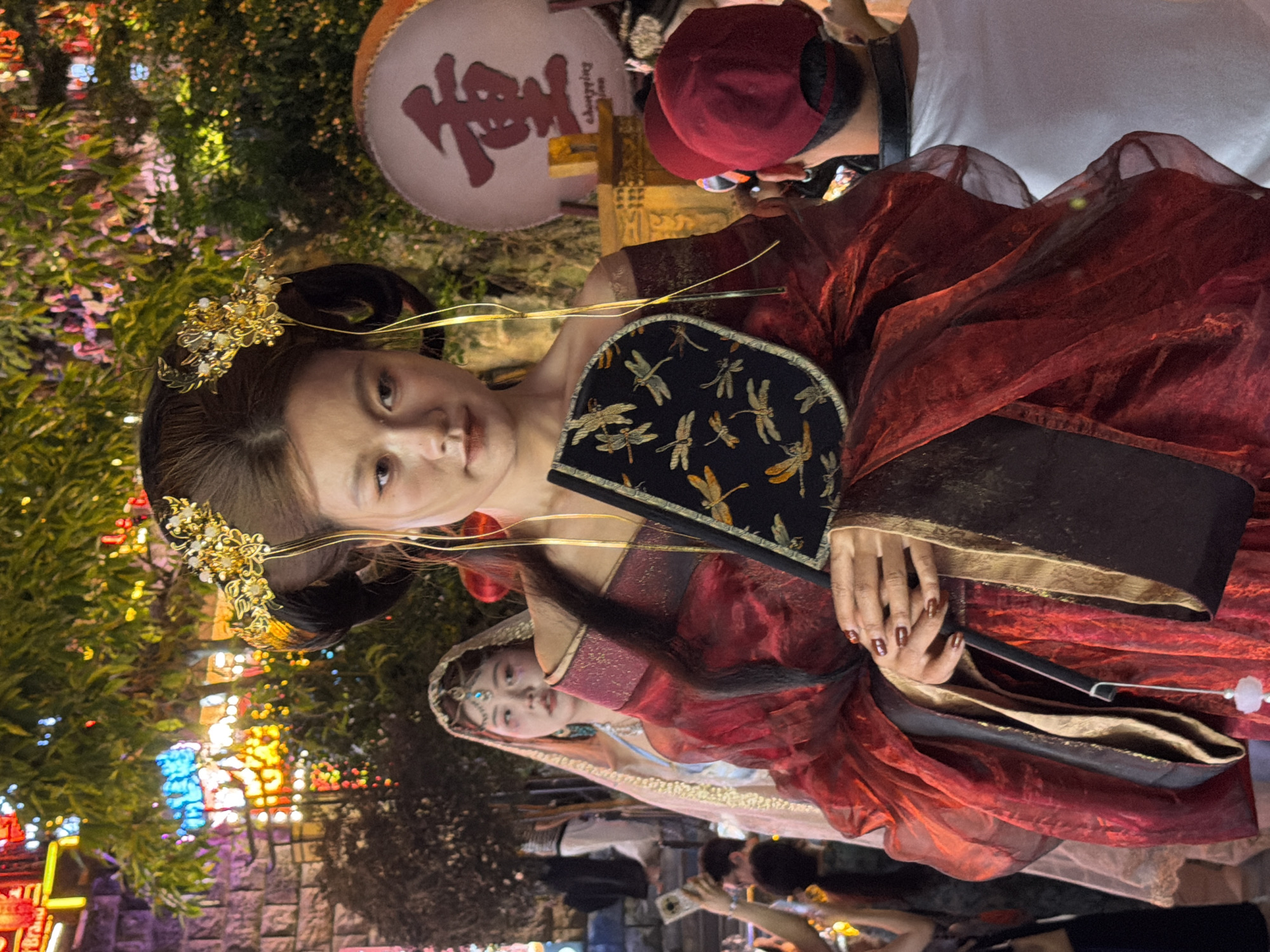 In Yuzhong District, China, a young woman in traditional red and gold Hanfu attire, adorned with elaborate golden hair ornaments, holds a black fan patterned with dragonflies. She is the central subject, facing forward with a neutral expression. To her left, another woman in a lighter colored, possibly pink, traditional outfit and veil stands partially visible. Behind the main subject, a large circular banner with a red Chinese character and the word "Chongqing" is displayed. The scene appears to be outdoors, likely at an event or festival, with colorful lights and foliage in the background. Several people are visible in the periphery, some taking pictures. The lighting suggests it is nighttime or dusk.