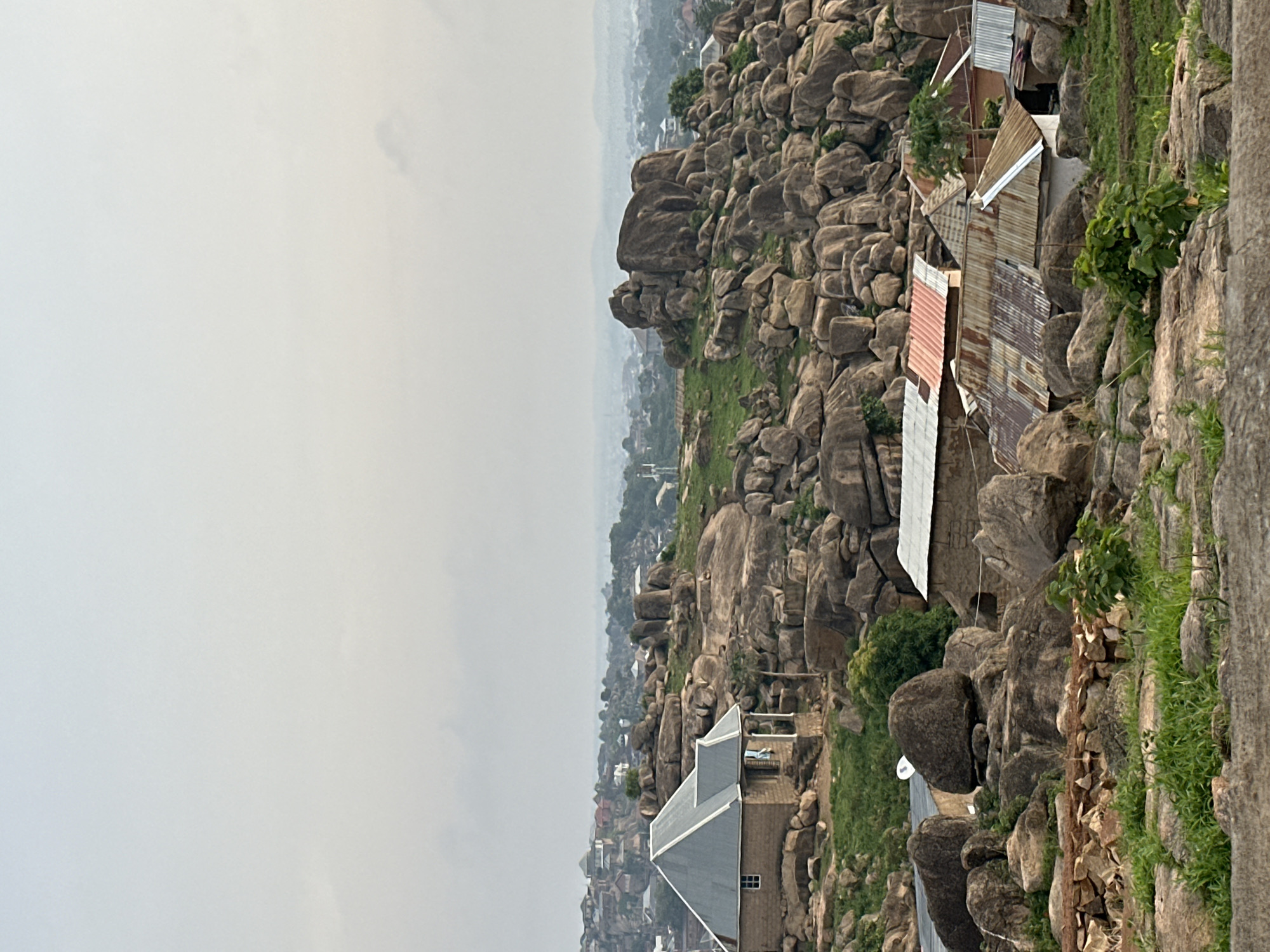 This is an elevated, wide-angle view of a rugged, rocky landscape under an overcast sky. The image captures a scene dominated by numerous large, rounded boulders and rock formations, characteristic of the Jos Plateau in Nigeria, where human settlements are intertwined with the natural terrain.
In the midground and foreground, the landscape is densely populated with grey-brown rocks, between which patches of green grass and sparse vegetation, including small trees and bushes, grow. Several houses are nestled amongst these formations. To the right, a cluster of more rustic homes features roofs made of various corrugated metals, some appearing rusty and weathered, others dull silver. To the left, a more substantial building with a neat grey pitched roof and a visible window stands out, suggesting a more modern construction.
Beyond the immediate rocky landscape, the view extends to a town or city, stretching into the distance. This urban sprawl is partially obscured by a soft, light grey atmospheric haze or fog, blending with the uniform, pale sky that dominates the upper portion of the scene.
The weather appears overcast, and the diffused lighting suggests it is daytime without direct sunlight. No people are visible, nor is any specific activity taking place. There is no visible text in the image.