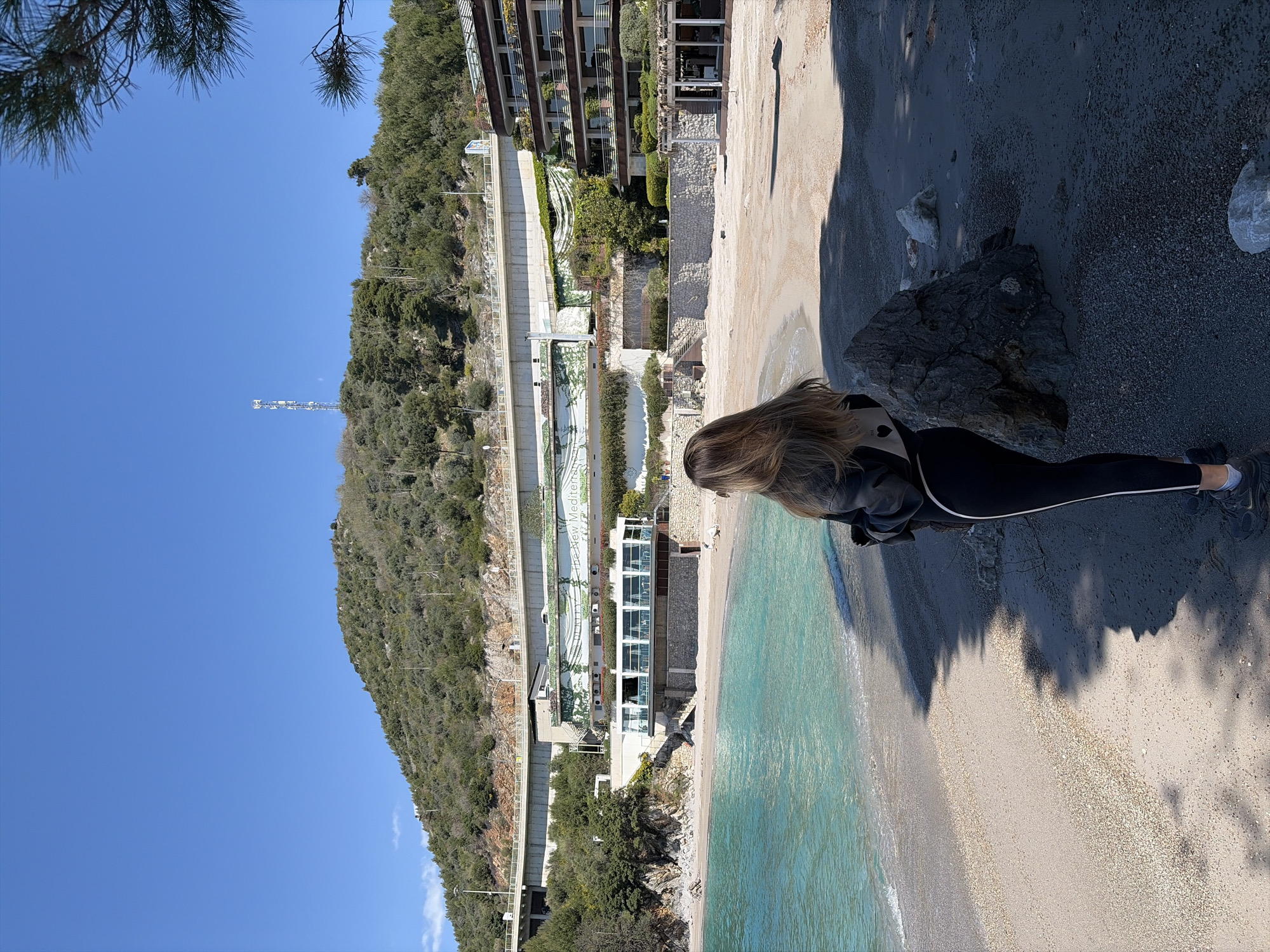 The image depicts a beach scene in Unknown, Unknown. A woman is standing on a dark, rocky area overlooking a sandy beach and clear turquoise water. The beach stretches along the coastline, bordered by a steep, forested hill. A road with retaining walls runs along the base of the hill, and several buildings are situated along the beach and hillside. The sky is clear and blue.