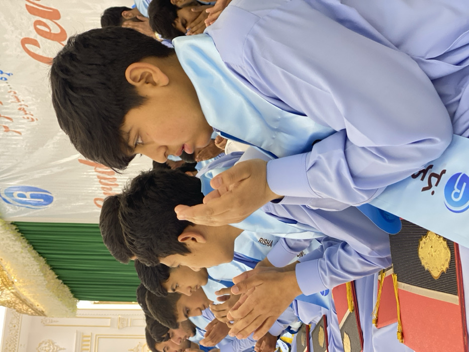 A group of young men in light blue uniforms and sashes stand in a row, their hands clasped together in prayer or reverence. They face forward, heads slightly bowed. Behind them, a banner with Arabic script is visible, along with green and white drapery. Each person holds a book, likely religious in nature, with golden ornamentation and red accents. The overall scene suggests a formal occasion, possibly a graduation or religious ceremony.