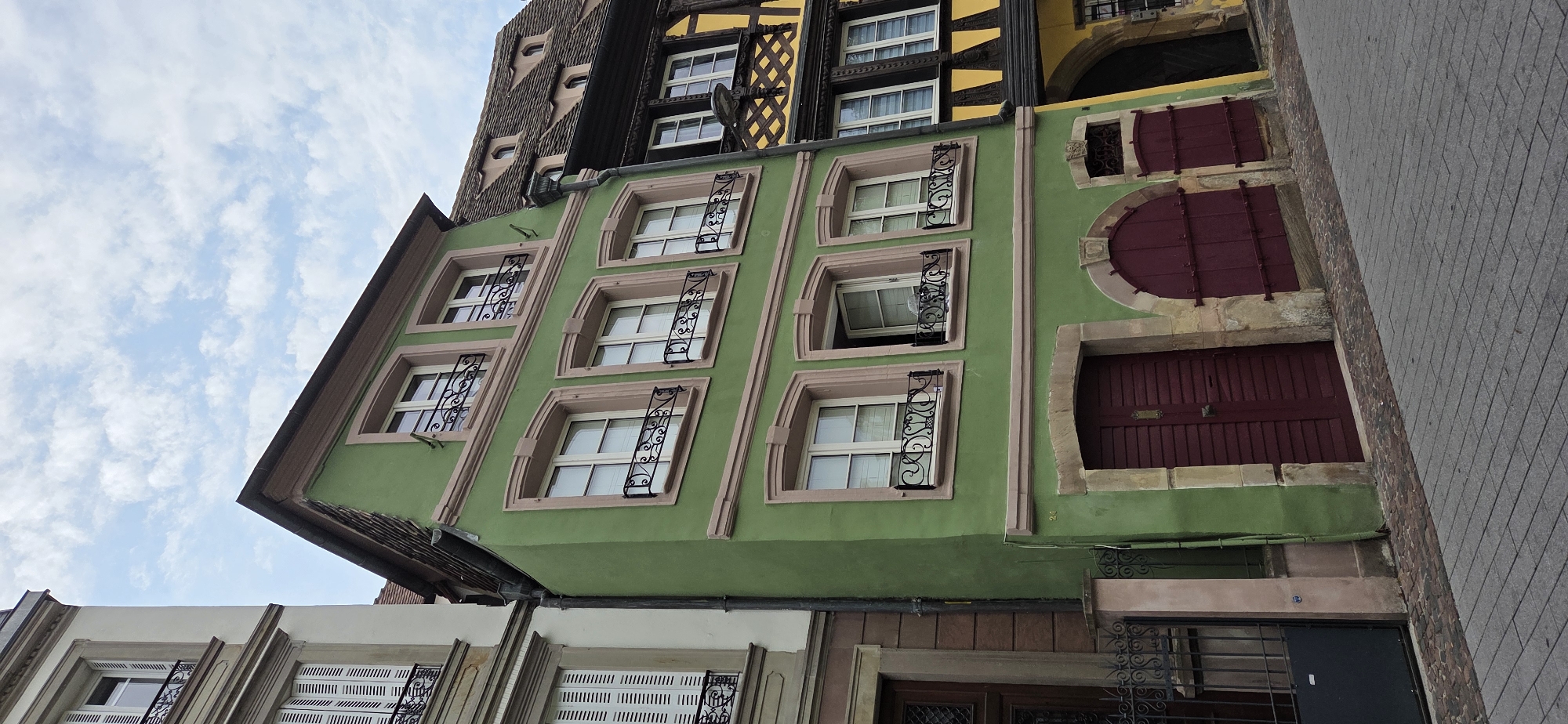 A tall, slender, lime green building with a triangular roof stands in the foreground, angled towards the viewer's right. It has three stories, each with two windows, except for the topmost story which has only one window. All windows are rectangular with white panes and black, ornate wrought-iron railings. The windows are framed by light pink, decorative moldings. The ground floor features two large, maroon, louvered doors set within arched stone frames. To the left of the green building is a sliver of a taller, cream-colored building with white louvered shutters on its windows. To the right of the green building is a yellow building with black timber framing. The buildings are situated on a street paved with cobblestones. The sky above is a light blue with scattered white clouds.