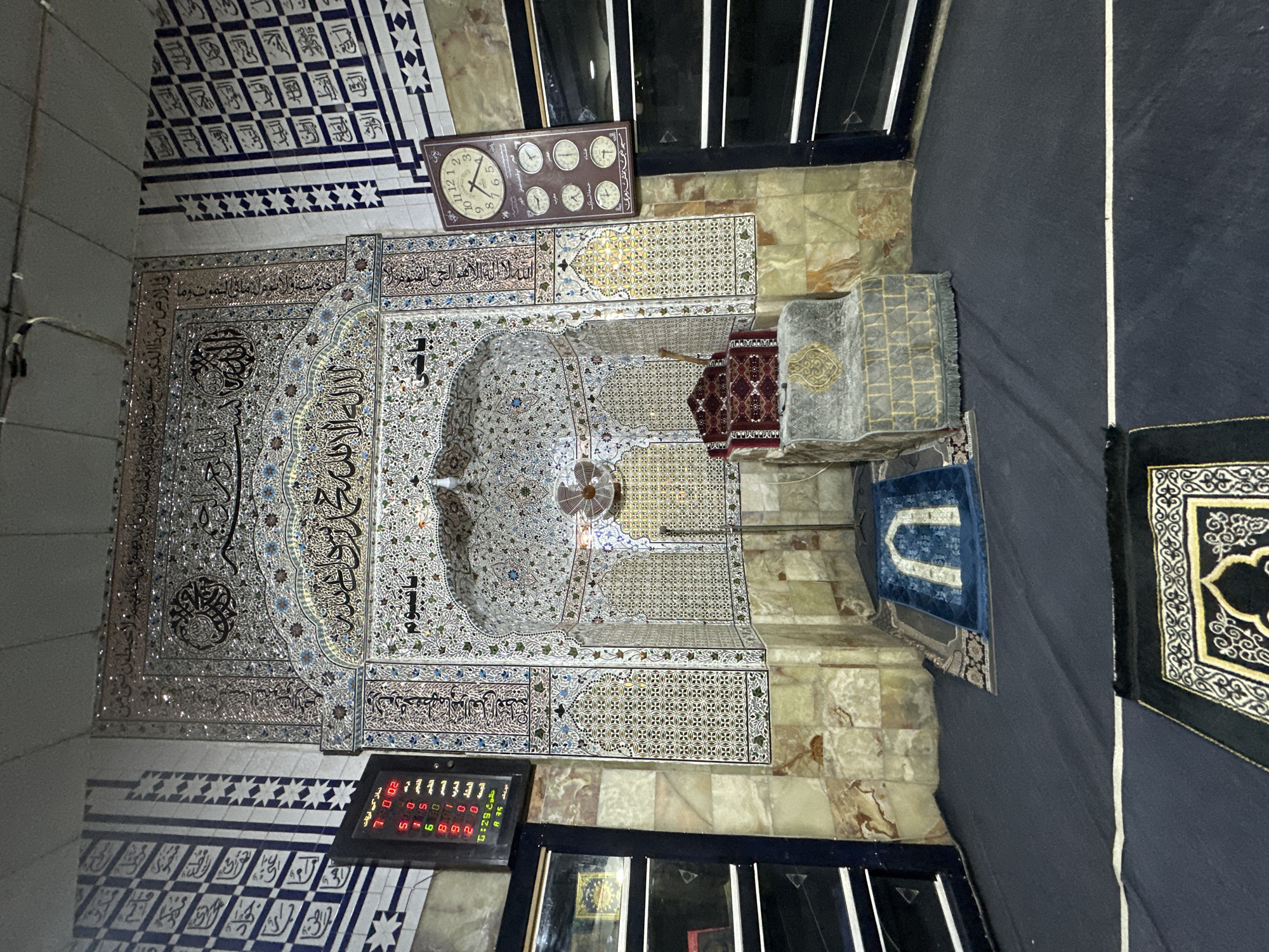The image depicts the interior of a mosque in Quetta City Tehsil, Pakistan. The focal point is the mihrab, an ornate niche indicating the direction of Mecca, adorned with intricate tilework featuring geometric patterns and Arabic calligraphy. Above the mihrab, a decorative arch is also covered in mosaic tiles and calligraphy. To the left of the mihrab, a digital prayer time display shows red LED numbers. To the right, a wall-mounted clock with multiple time zones is visible, along with a fan mounted on the wall behind the mihrab. In front of the mihrab, a raised, cushioned platform and a smaller, more ornate platform, possibly a minbar, are present. Two prayer rugs are laid out on the dark carpeted floor. The walls are decorated with patterned tiles and what appears to be marble. The overall impression is one of religious reverence and decorative artistry within a prayer space.