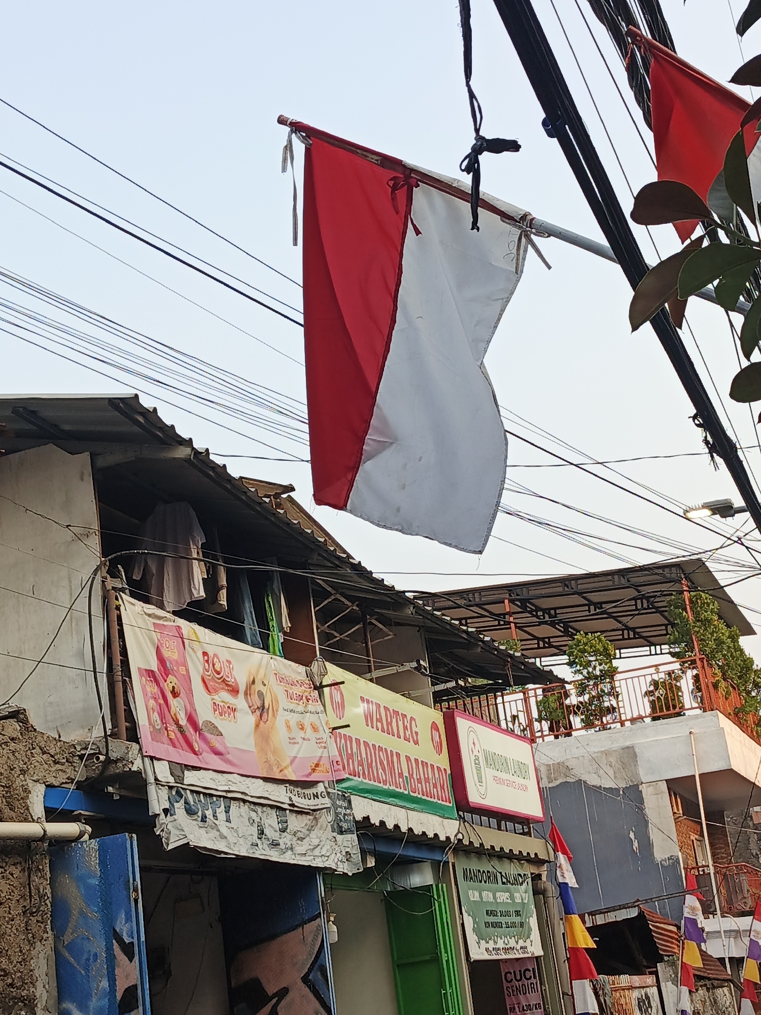 A red and white Indonesian flag flies from a pole in front of a multi-story building in South Jakarta, Indonesia. The building appears to be a mix of residential and commercial spaces. On the lower levels, several banners are displayed, including one for "BOLT PUPPY" dog food and another for "WARTEG KHASIRMA BAHARI". Above these banners, clothes are hung out to dry on a line. The upper floors show balconies with plants and railings. A street light is visible amongst a tangle of electrical wires overhead. The scene suggests a bustling urban environment with signs of daily life.