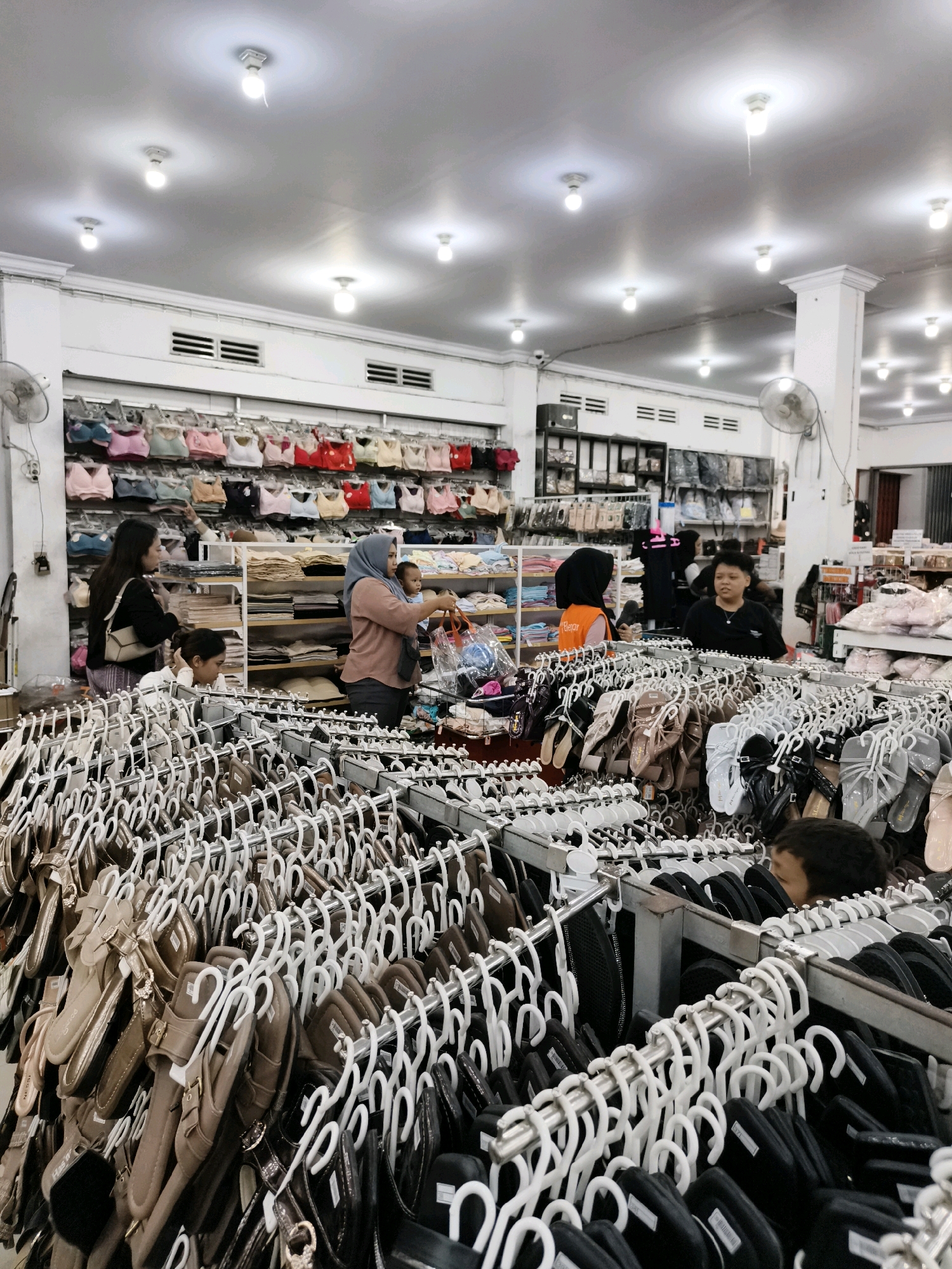 The image depicts the interior of a retail store, likely a clothing and accessories shop, located in the city of Ketapang, Indonesia. The store is brightly lit by numerous ceiling lights. The foreground is dominated by racks of hanging footwear, primarily sandals and flip-flops, displayed on white hangers. These are predominantly in neutral and metallic tones like brown, beige, gold, and black. In the midground, several individuals are visible. On the left, a woman in a black jacket and striped pants is looking at items. Next to her, another woman wearing a headscarf and a salmon-colored top is holding a baby and a shopping bag. Further back, a person in an orange vest, possibly an employee, is interacting with another individual. The background reveals shelves stocked with more merchandise, including what appear to be bras or bralettes, bags, and other apparel. The overall environment suggests a busy retail setting.