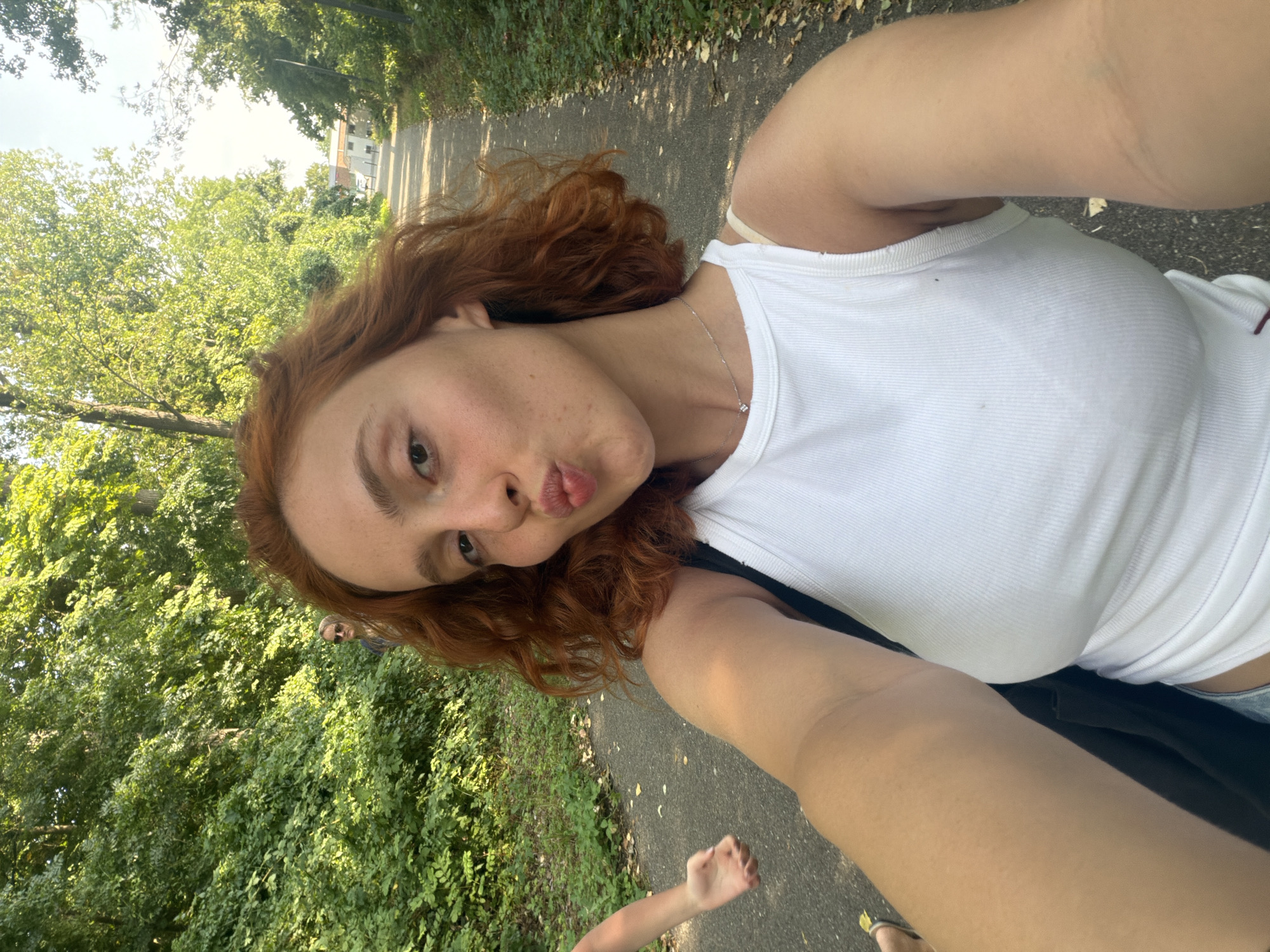 A young woman with auburn hair and freckles is taking a selfie outdoors. She is wearing a white tank top and a necklace. Her lips are pursed as if she is about to kiss. Her left arm is extended, holding the camera. To her left, a person's arm and torso are visible, with their arm outstretched. Further in the background, another person wearing sunglasses is standing amongst lush green trees. A paved path stretches into the distance, flanked by vegetation on both sides. Buildings are visible in the far distance. The scene appears to be a park or a nature trail within a semi-urban environment, potentially in Illkirch-Graffenstaden, France, given the presence of paved paths and buildings juxtaposed with dense greenery.