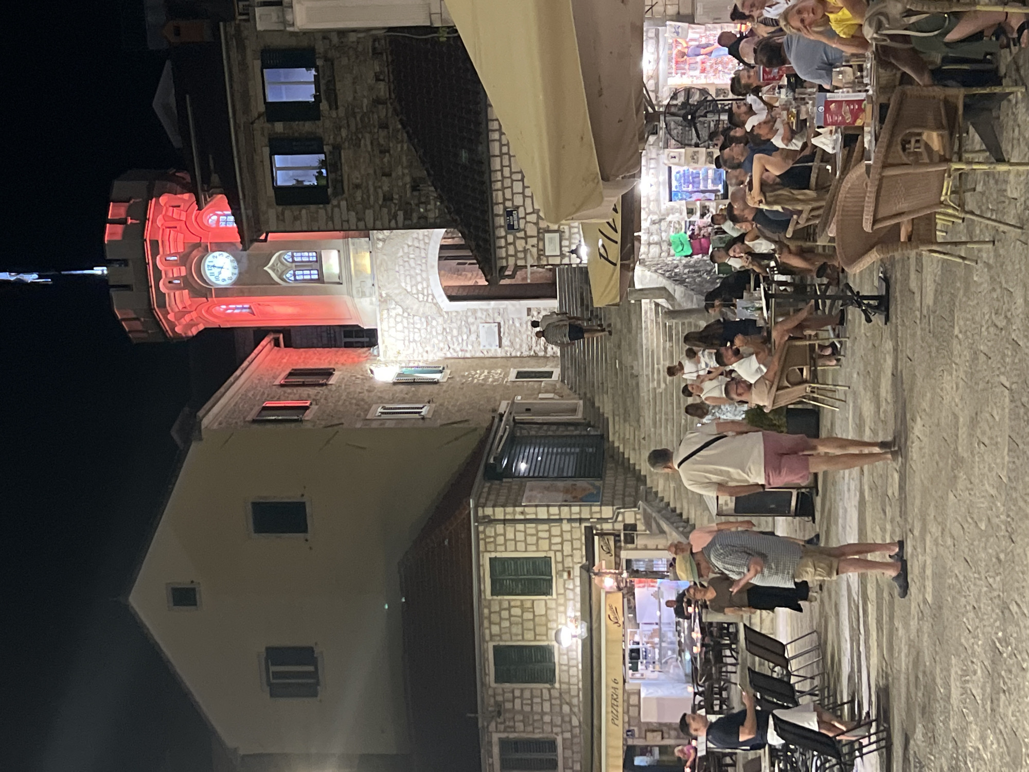 The image depicts a lively evening scene in the Old Town of Herceg Novi, Montenegro. A prominent clock tower, illuminated in red, stands tall in the background. In the foreground, people are seated at tables outside a pizzeria, engaged in conversation and dining. The cobblestone street and stone buildings suggest a historic urban setting. The overall atmosphere is one of a bustling European city square at night.
