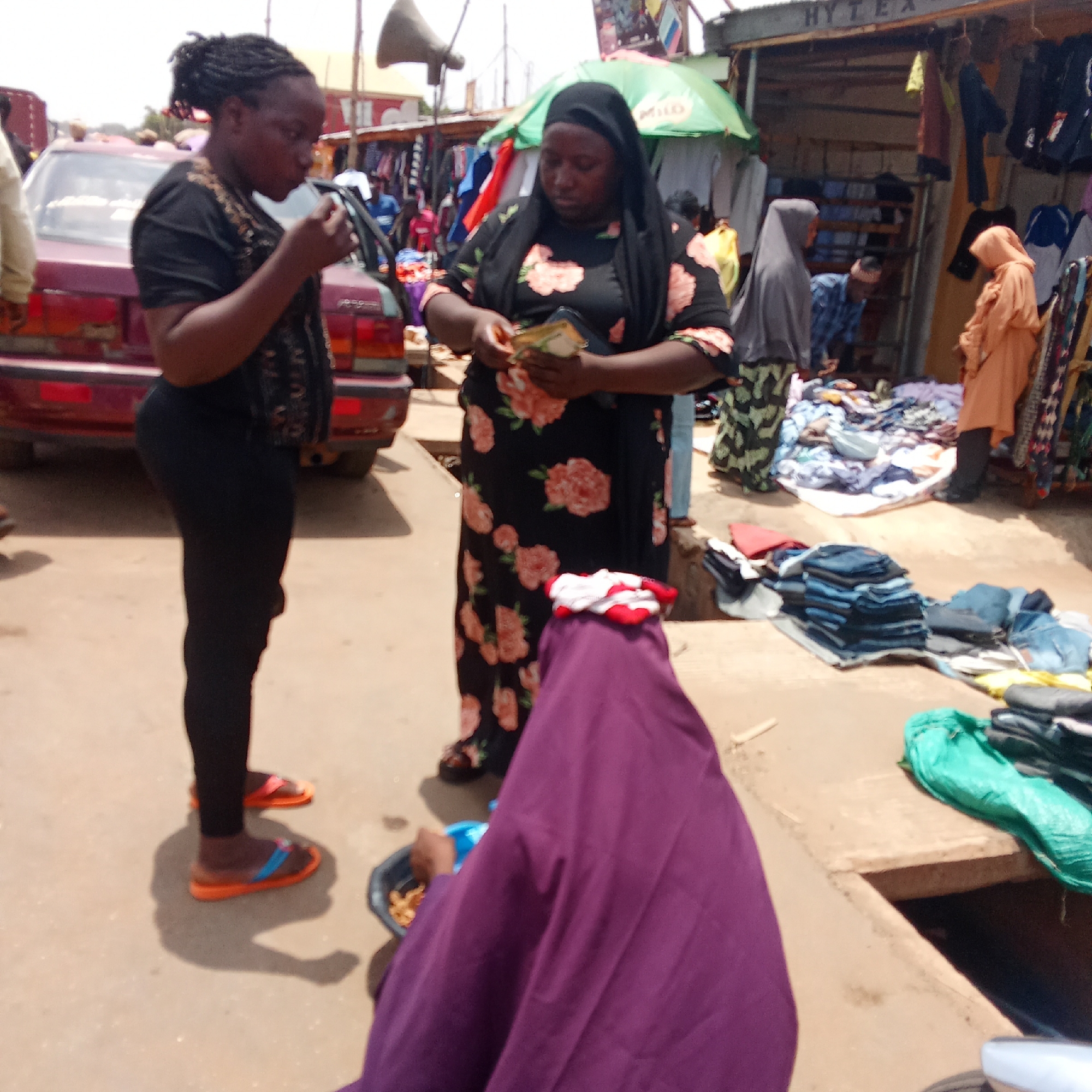 The image shows a street scene in Akwanga, Nigeria. In the foreground, a woman in a black outfit and orange sandals is eating. Next to her, a woman in a black floral dress and a black hijab counts money. A person in a purple garment is sitting on the ground, possibly selling something in a container.
In the background, there is a maroon car and several market stalls selling clothes. The stall furthest from the camera has a sign that reads "HYTEX". Other people are visible in the background, including women wearing hijabs and a man in a plaid shirt. The scene appears to be a bustling marketplace.