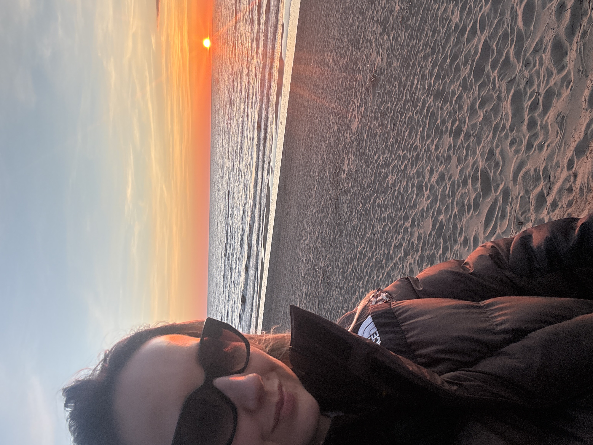 A person wearing a black puffer jacket and sunglasses is seen in a selfie taken at a beach at sunset. The beach is sandy and the sea reflects the sunset colors. The sky is transitioning from blue to orange near the horizon. The location is Garupe, Latvia.