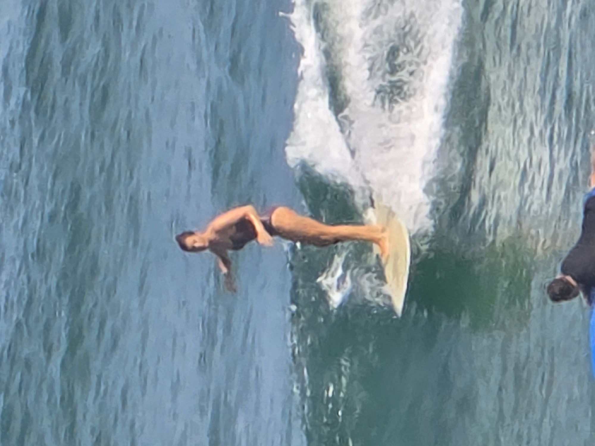 A man is surfing on a board in the sea off the coast of Tel Aviv, Israel. He is wearing dark swimwear and is riding a wave. The water is turbulent, creating a spray behind the surfer. A person is partially visible on the right side of the image, likely observing the surfer. The scene is taken during daylight.