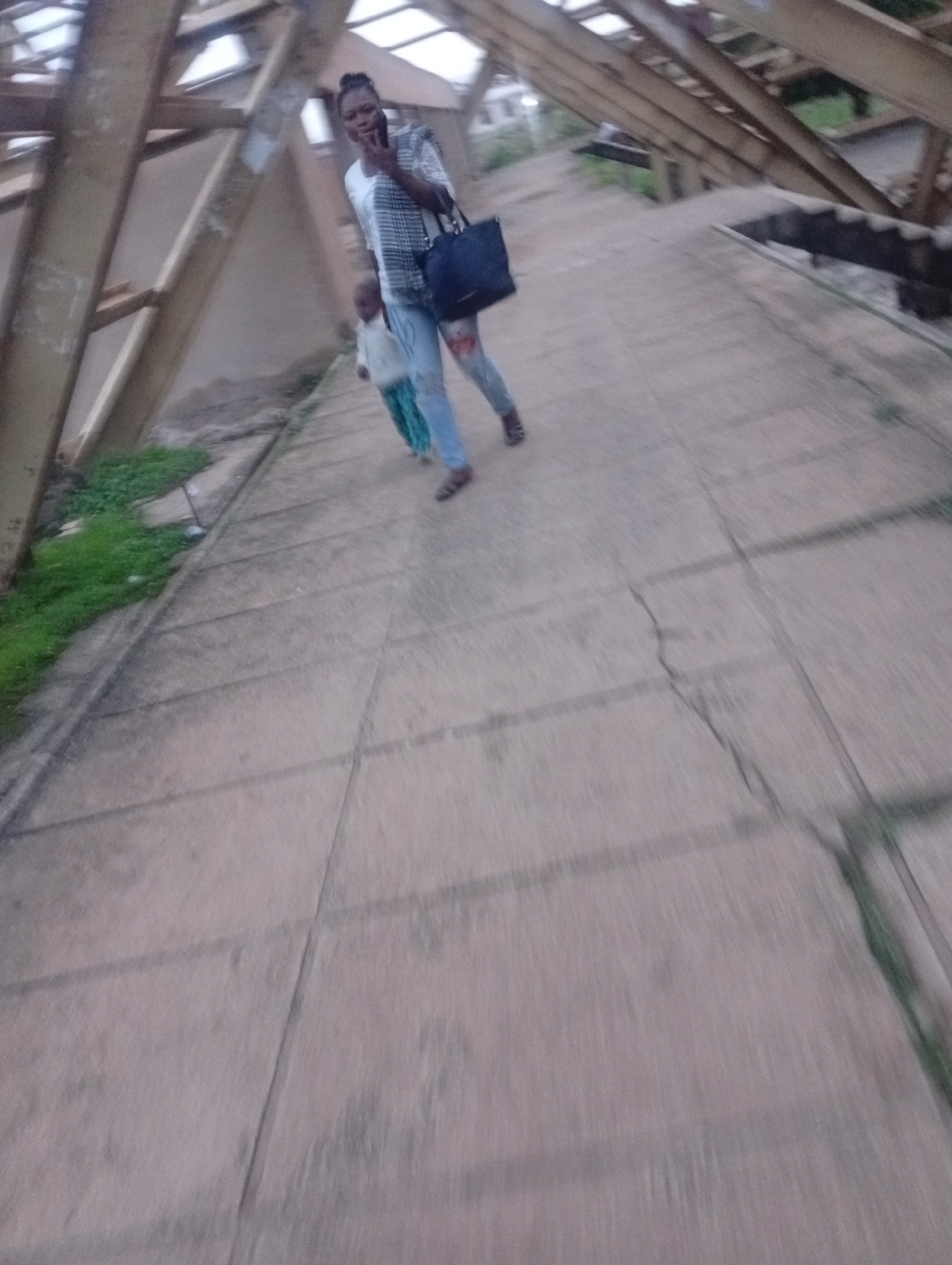 In an outdoor, open-air structure with a sloped, translucent roof and prominent wooden beams, a Black woman is walking down a tiled pathway. She is wearing a plaid shirt over a white t-shirt and blue jeans, and carries a dark bag over her left shoulder. To her right and slightly behind her, a child in a green and white patterned garment is walking in the same direction. The pathway is cracked and weathered, with some patches of green grass visible on the left side. The overall impression is one of casual movement within a somewhat dilapidated or under-construction urban environment in Jos, Nigeria.
