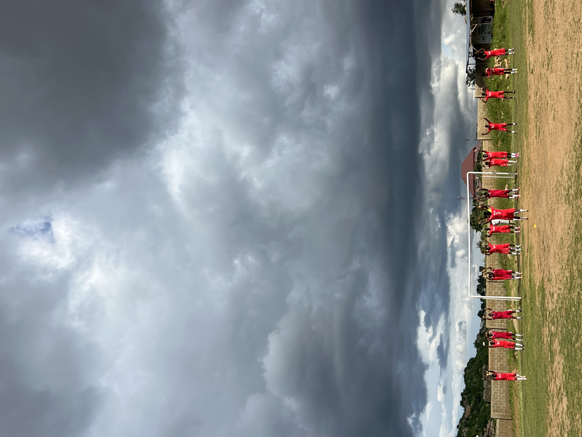 This image shows a group of young male soccer players in red uniforms on a grassy field, standing in front of a goal. They are all raising their arms in the air. The sky above is filled with dark, ominous storm clouds. In the background, there are buildings, some with corrugated iron roofs, and a rocky hillside dotted with sparse vegetation. The scene appears to be an outdoor soccer training session on a cloudy day, possibly in Jos, Nigeria.