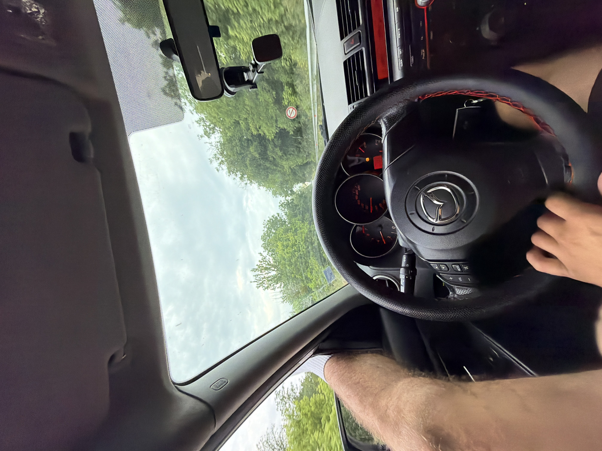 An interior view from a moving car captures the driver's perspective in Gorna Studena, Bulgaria. A hand rests on the bottom of a black Mazda steering wheel, which features red stitching and integrated controls. Car keys hang from the ignition behind the steering wheel. To the left, a leg with a white sock is propped against the dashboard, adjacent to the A-pillar labeled "AIRBAG." The instrument cluster displays red-lit gauges and a red warning indicator light. A center console with red-accented controls and a rearview mirror with an attached phone holder are visible. Beyond the windshield, an overcast sky is visible above a road flanked by dense green trees. A white circular road sign with a red border and a diagonal line through two opposing arrows (End of no overtaking) is visible on the right side of the road.