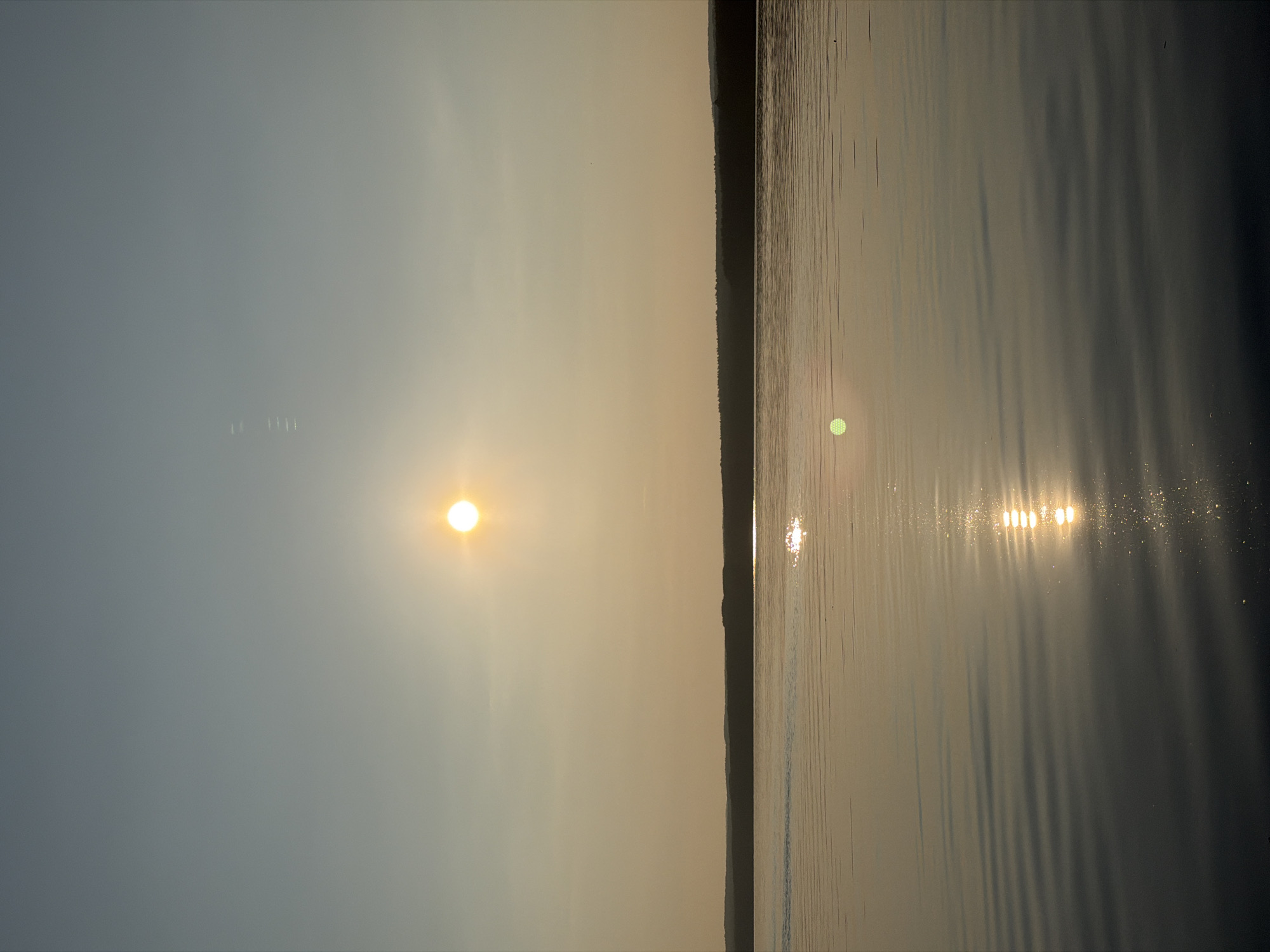 The image depicts a serene sunset over the Kama River in Chaikovsky, Russia. The sun, a bright orb, is positioned high in the sky, casting a warm, golden glow. The sky transitions from a pale blue at the top to a soft, hazy yellow near the horizon. A dark, silhouetted treeline marks the distant shore. The water's surface is calm, reflecting the sun's light in shimmering patterns of gold and white. Gentle ripples create a textured effect on the water, adding depth to the scene. There are no people or specific objects visible in the foreground or midground. The overall atmosphere is peaceful and tranquil.