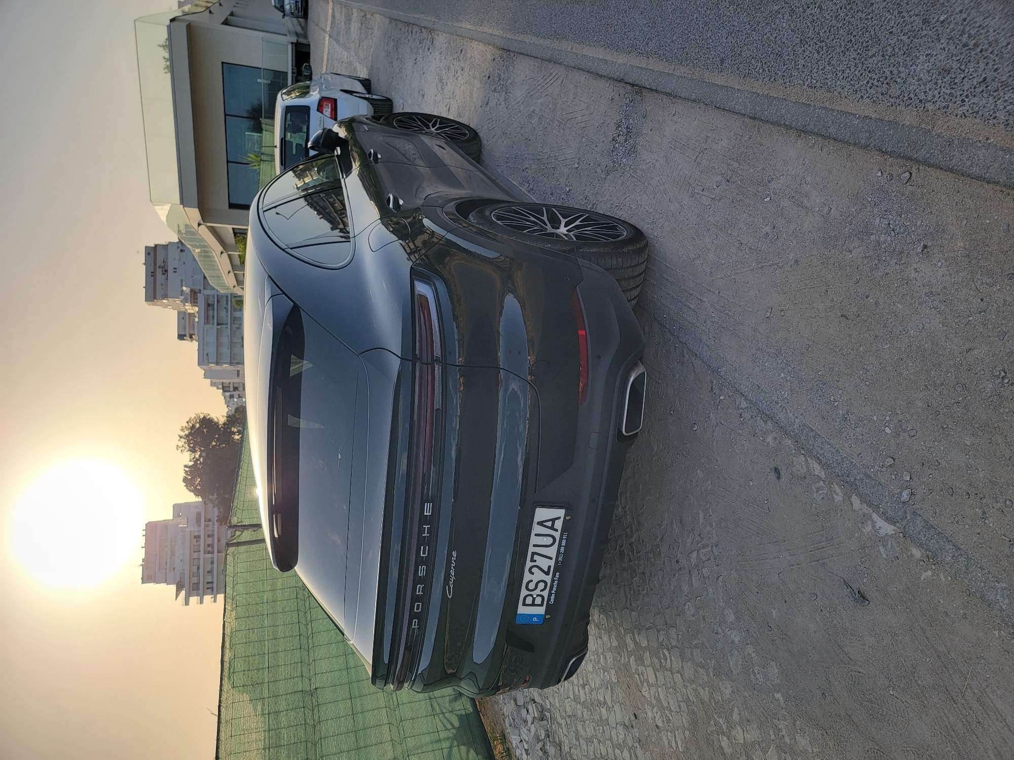 The image depicts the rear of a dark grey Porsche Cayenne parked on a dirt and gravel surface. The car has a Portuguese license plate that reads "BS 27 UA". In the background, a modern building with a glass railing and large windows is visible, along with several apartment buildings. The setting sun casts a warm glow, creating lens flare. A green construction fence is present on the left side of the frame. The scene appears to be in Quarteira, Portugal, likely near a construction site or undeveloped area adjacent to a road.