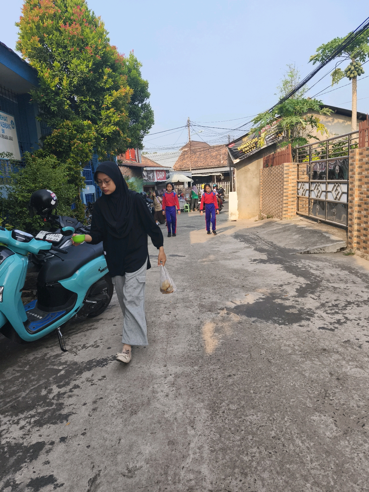 In Palembang, Indonesia, a young woman wearing a black hijab and grey pants walks down a sun-drenched street, holding a plastic bag of food in her right hand and a green bowl in her left. She is near a turquoise scooter parked on the right side of the path. In the background, two girls in red shirts and blue pants walk away from the camera, and several buildings line the street. The ground is paved with concrete, showing signs of water stains and a few scattered yellow leaves. The sky above is a clear, pale blue.