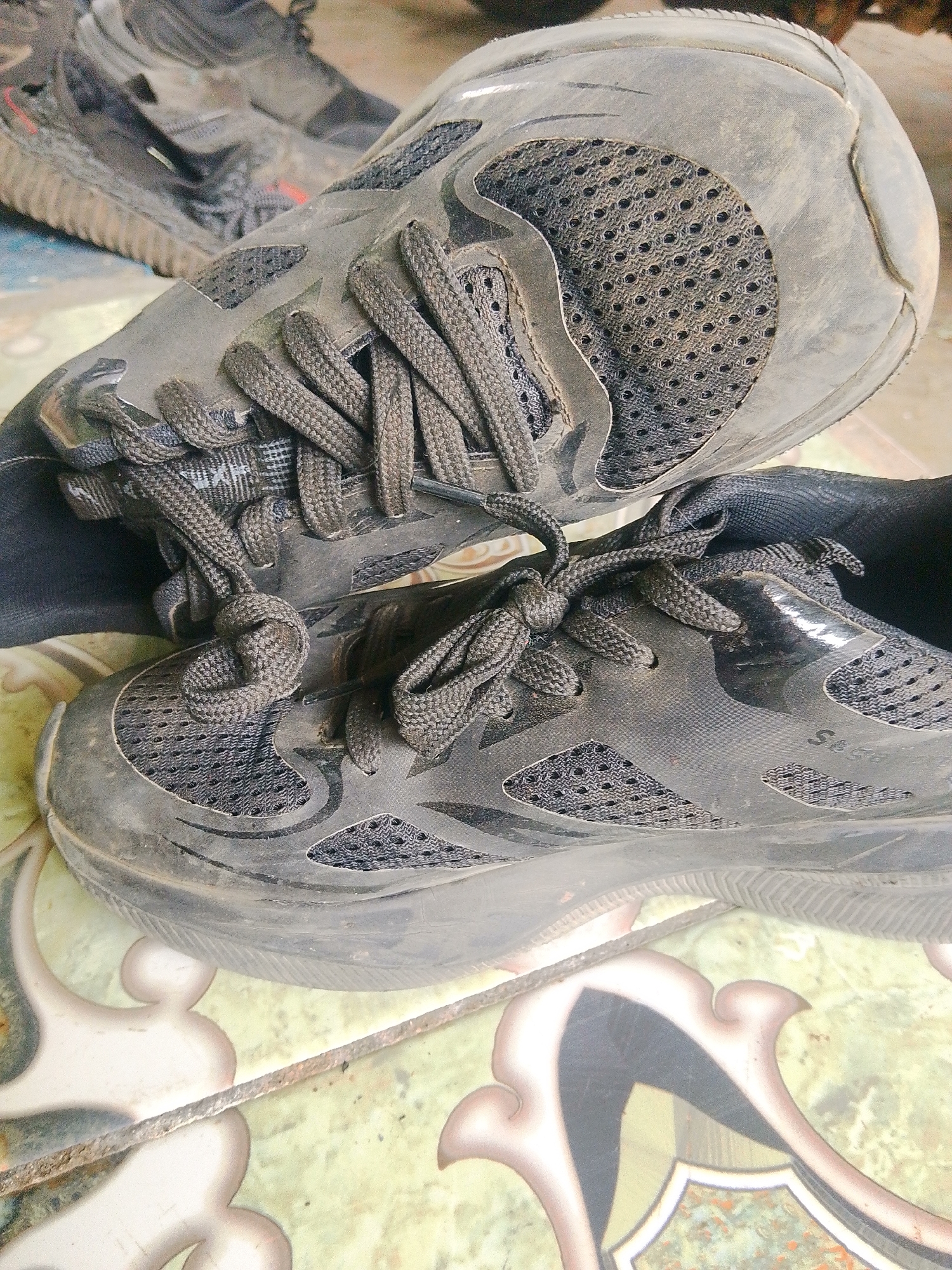 This is a close-up, high-angle shot of two pairs of dusty, dark-colored athletic shoes resting on a tiled floor. The shoes are worn and appear to have been used outdoors, as indicated by the dust and dirt covering their surfaces. The foreground shoe is a lace-up sneaker with a mesh upper and a thick sole. The laces are tied in a knot. Behind it, another pair of similar shoes is partially visible, one shoe angled upwards, obscuring part of the shoe in front. The tiled floor has an ornate pattern with light green and cream colors, featuring decorative swirls and a central shield-like design. The overall scene suggests a casual, possibly utilitarian, setting. The location is identified as Barabai, Indonesia.