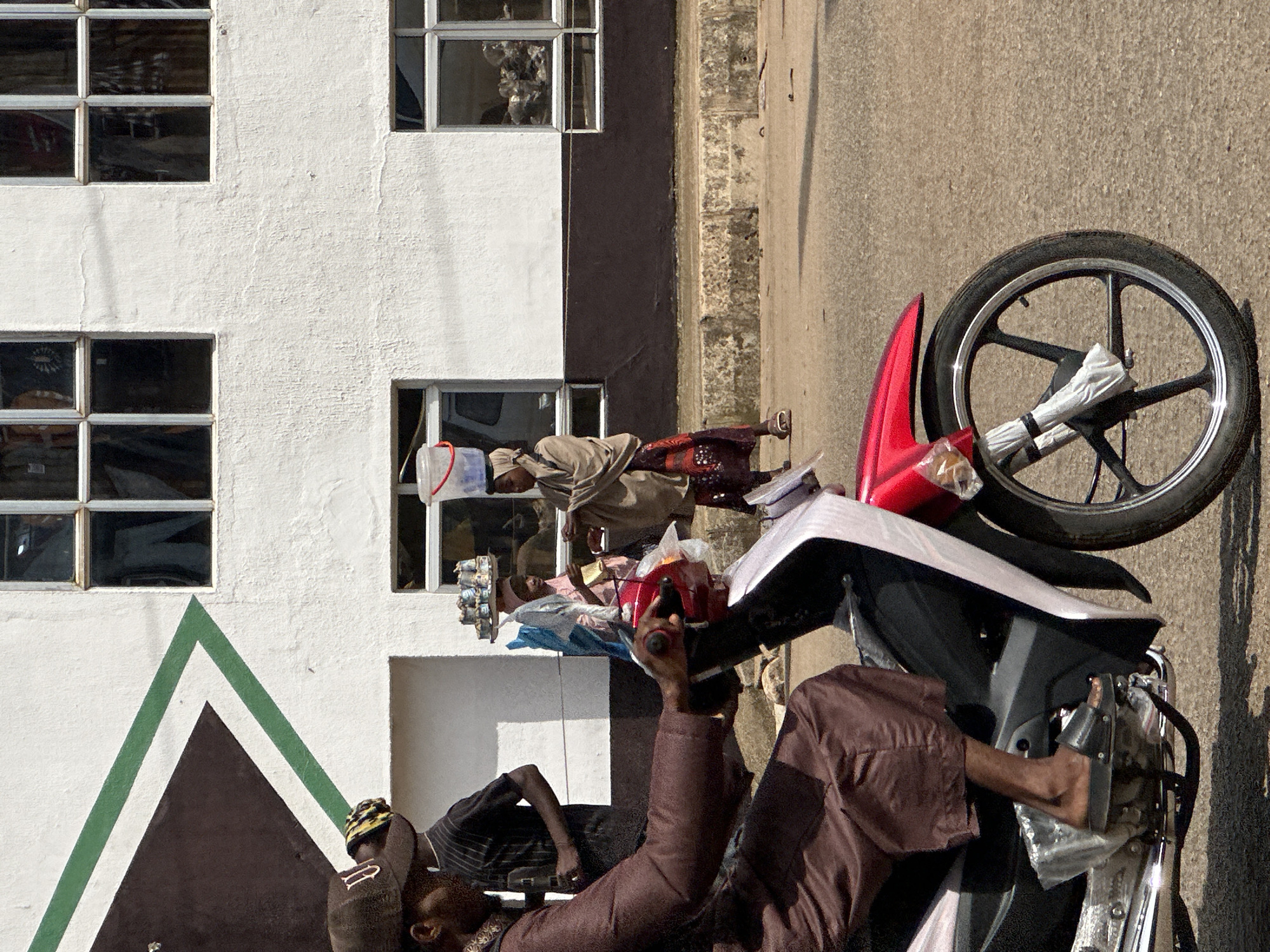 The image depicts a street scene in Bukuru, Nigeria, focusing on a man riding a red motorcycle in the foreground. He is wearing a brown robe and a brown cap. Behind him, several other individuals are visible. To the left, a person in a black and white striped shirt stands near a building with a distinctive triangular design featuring green and brown accents. Further back, two women are walking. One woman, wearing a pink headscarf, carries a tray of goods on her head. The other woman, in a beige headscarf and patterned skirt, also carries a bucket on her head. The background is dominated by a white building with multiple windows. The street surface is asphalt, and a concrete curb is visible along the side of the road. The overall impression is a snapshot of daily life in a Nigerian urban environment.