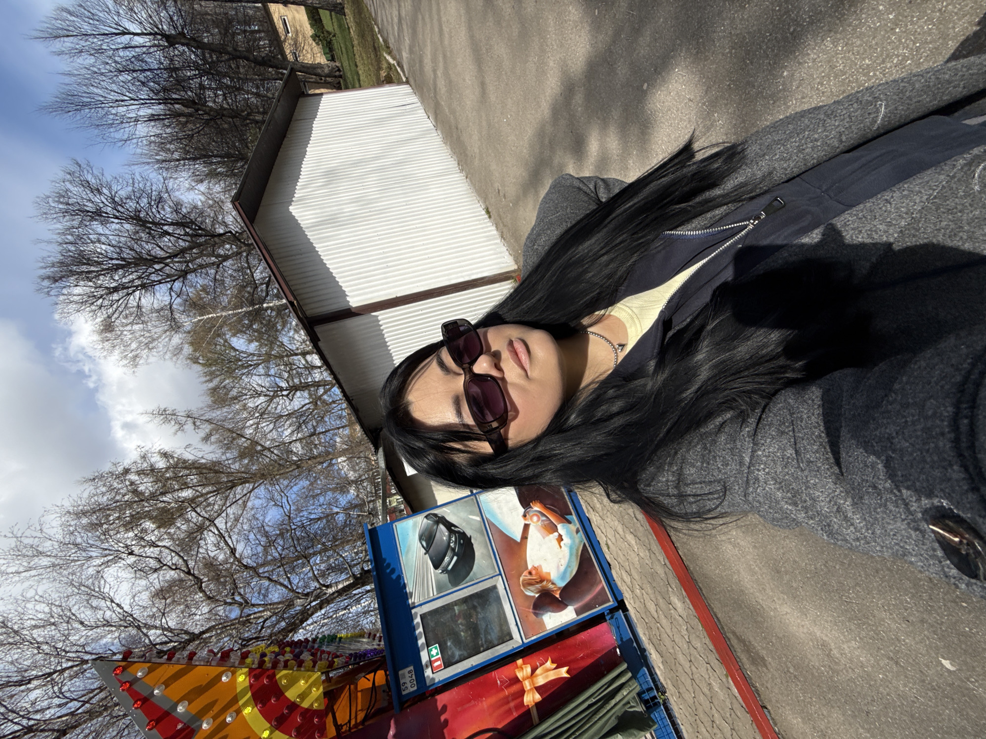 A woman takes a selfie outdoors on a sunny day in what appears to be a park or amusement area in Perm, Russia. She is positioned off-center to the right, looking towards the camera with sunglasses on. She has long, dark hair, light-colored lips, and is wearing a light yellow top under a dark blue zipped jacket and a grey coat. A silver chain necklace with a small pendant is visible around her neck.
Behind her, to the viewer's left, is a colorful structure adorned with numerous red, yellow, and clear lights, suggesting part of a fairground ride. To her right, a blue vending machine or display unit stands prominently. It features various images: a black car (resembling a Nissan Juke or similar crossover) at the top, and a depiction of a woman with blonde hair looking up towards a light source on the bottom right. A screen or panel on the bottom left displays a red cross and a green plus sign, along with the numbers "59" and "0048".
In the background, a white corrugated metal building with a dark roofline is visible. Beyond that, numerous bare trees with intricate branches suggest the setting is either late autumn or early spring. The ground is paved with asphalt or concrete, featuring a distinct red curb line. The bright sunlight casts long, clear shadows, indicating either mid-morning or late afternoon. The sky is partially cloudy with patches of blue visible.