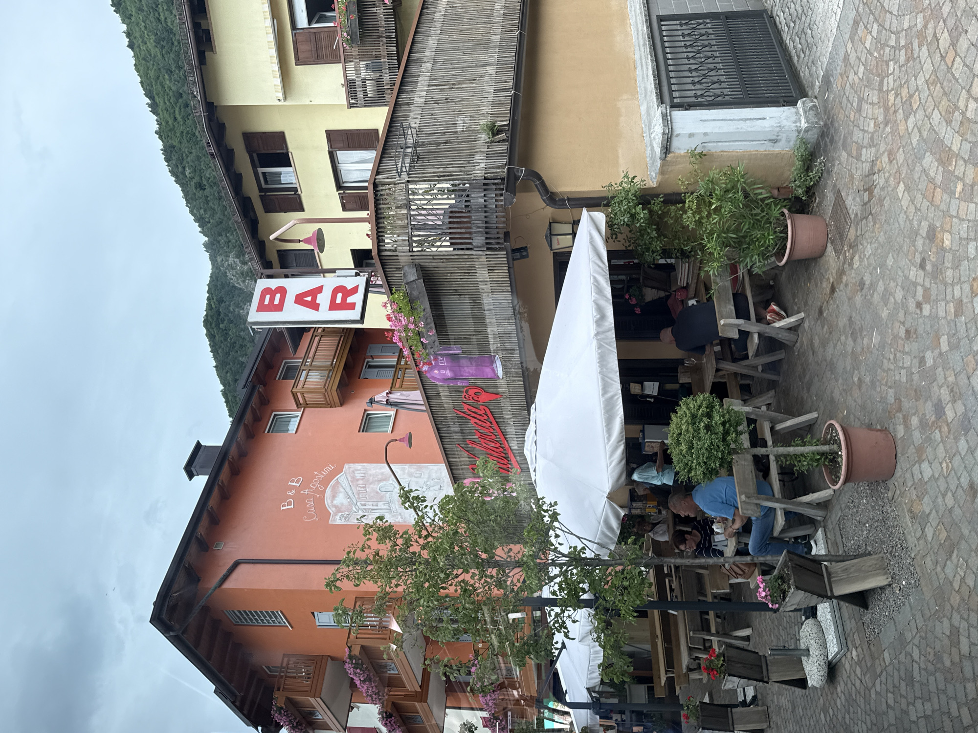 In the city of Fai della Paganella, Italy, an outdoor seating area of a bar and B&B is depicted. The establishment features an orange building on the left with the inscription "B&B Casa Agostini" and a mural. To its right is a yellow building. A prominent red "BAR" sign hangs vertically. The area is furnished with wooden tables and chairs, some covered by white umbrellas. Several patrons are seated at the tables. A man in a blue shirt and jeans sits at one table, while another man with a shaved head is seen from behind at a different table. A woman with dark hair and glasses sits with the man in the blue shirt. A tree stands near the seating area, and potted plants are strategically placed throughout the scene. The ground is paved with cobblestones. The sky is overcast, and a forested mountain slope is visible in the background.