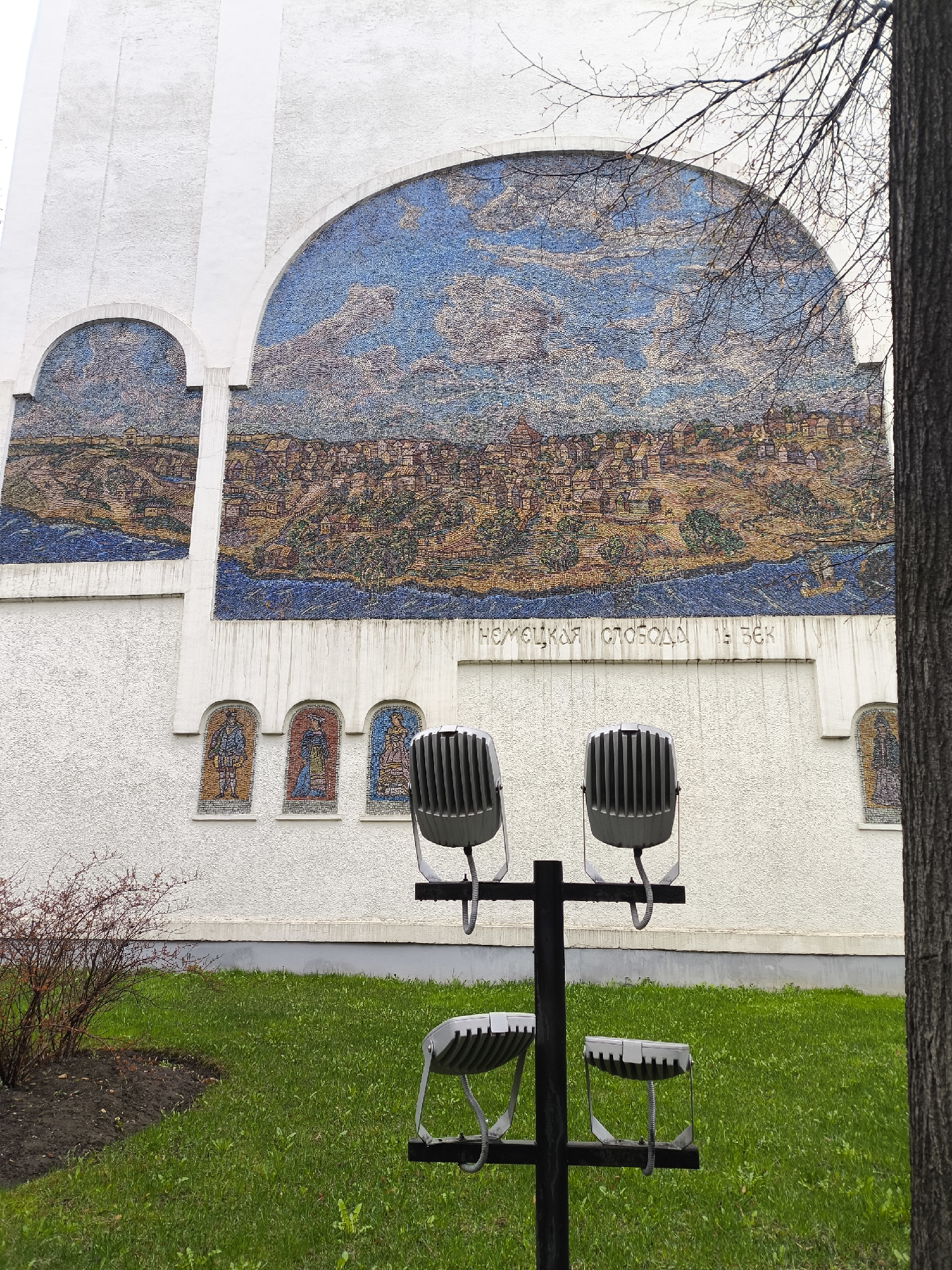 A daytime shot shows a mosaic art piece on a wall, likely in Moscow, Russia, judging by the Cyrillic script "НЕMEЦКАЯ CЛОБOДA 16 BEK" visible below the artwork. The mosaic depicts a townscape under a sky with scattered clouds. Below the main artwork are smaller, arched mosaics depicting figures. In the foreground, there's a metal lighting fixture mounted on a pole, and a small patch of grass and a bush at the base of the wall. A tree trunk is visible on the right side of the frame, obscuring part of the mural. The building has a textured, off-white facade.