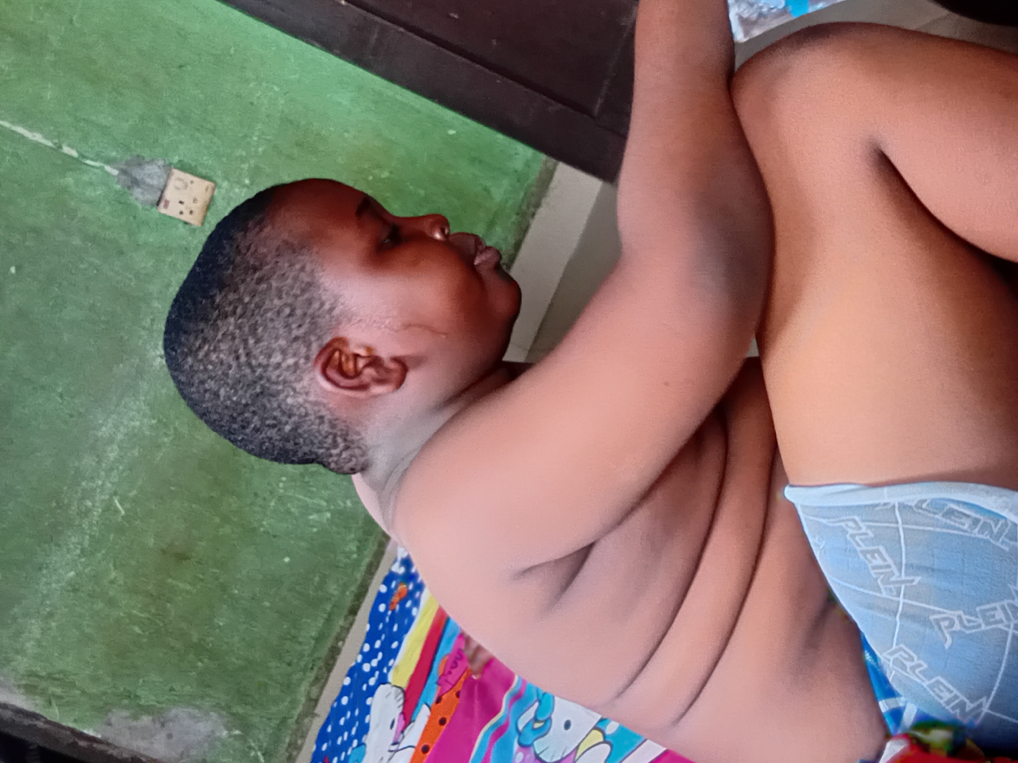 This is a photograph of a young child lying on their side. The child has dark skin and short, dark hair. They are wearing light blue underwear with a repeating white "PLEIN" logo. The child appears to be resting on a colorful blanket, which has a pattern of white cartoon characters and stripes in various colors including blue, pink, yellow, and orange. To the left of the child is a green surface, possibly a floor or mat, with a white electrical outlet visible on it. In the background, there is a dark brown wooden structure, possibly a table or piece of furniture. The overall scene suggests an indoor or semi-outdoor domestic setting in Nkpor, Nigeria. The lighting appears to be natural, indicating it could be daytime. There are no obvious emotional cues from the child's expression, as their face is partially obscured.