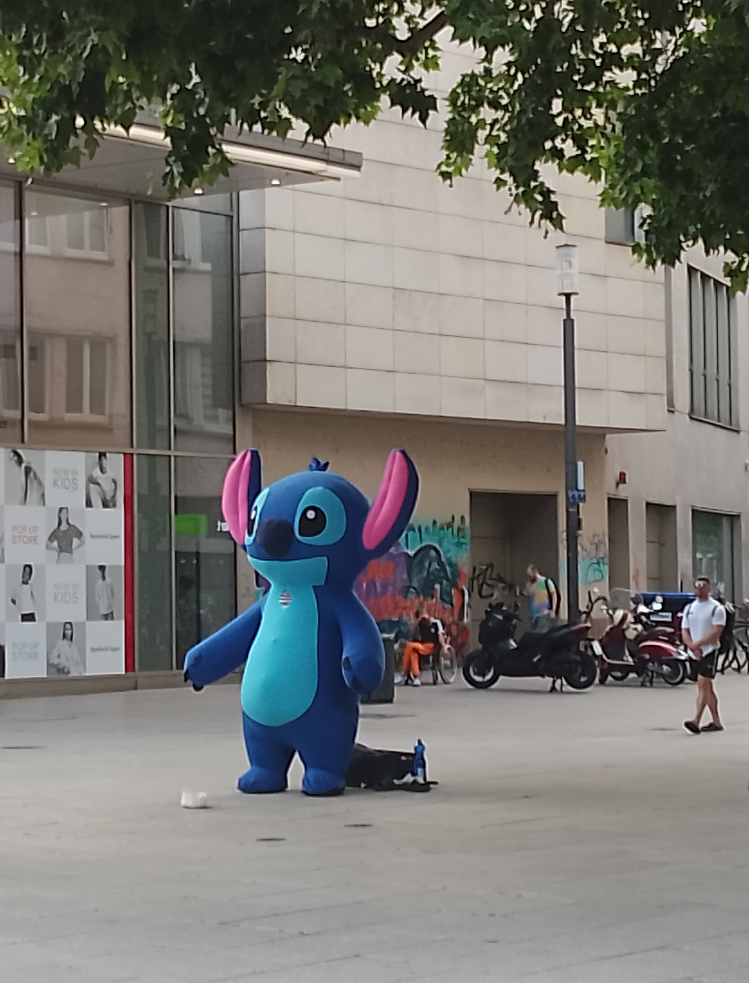 A blue Stitch costume character stands on a paved urban plaza. The character is facing towards the right of the frame and appears to be interacting with something on the ground. In the background, there is a modern building with large glass windows, a street lamp, and several parked scooters and motorcycles. A person in a white t-shirt and black shorts is standing to the right of the frame, facing left. The plaza is empty of other people except for the individual mentioned. The sky is not visible due to the overhanging trees.