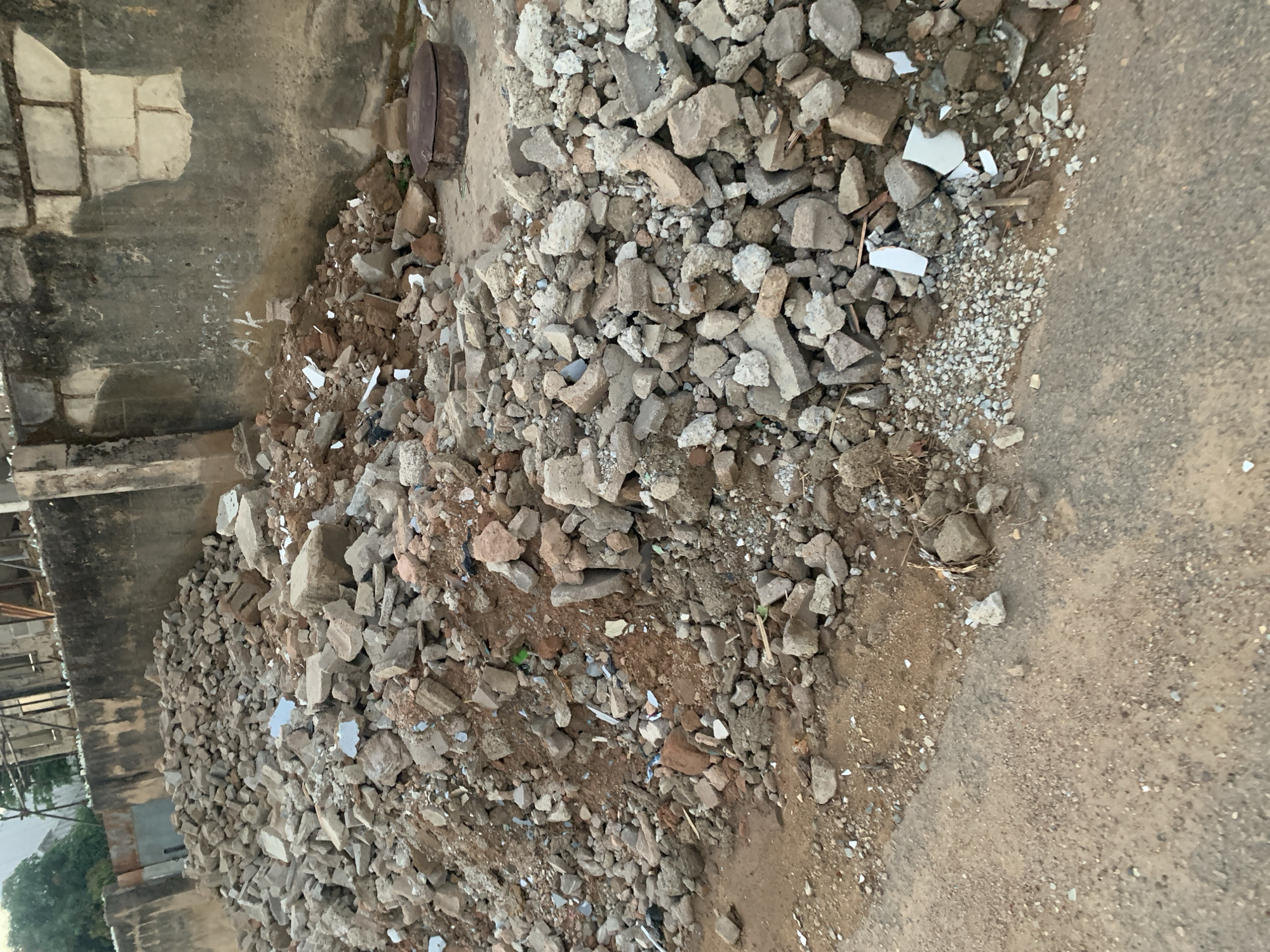 A pile of rubble consisting of broken concrete, bricks, and other debris sits against a weathered, concrete wall and spills onto an asphalt road in Kabong, Nigeria. The rubble is composed of varying sizes of fragments. The asphalt road shows signs of wear. The wall is stained. Vegetation is partially visible in the background.