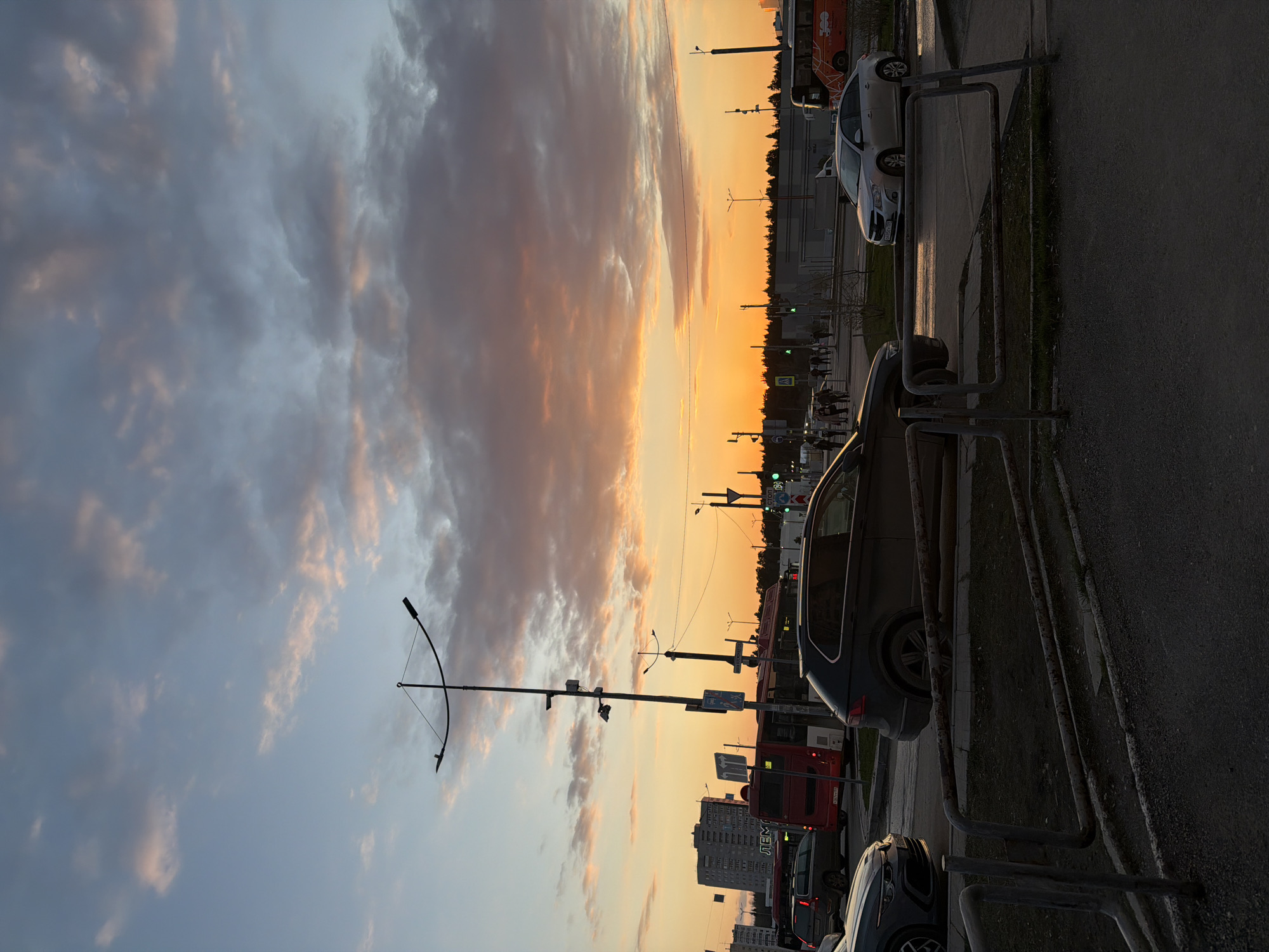 The image captures an urban street scene during what appears to be sunset or sunrise, evidenced by the vibrant colors in the sky. The horizon, positioned approximately one-third up from the bottom of the frame, glows with an intense orange-yellow hue that gradually transitions into softer blue and grey clouds occupying the upper two-thirds. The sky is partly cloudy, with scattered formations reflecting the warm light.
In the foreground, a paved area, likely a sidewalk or roadside, is separated from the street by weathered metal barriers. Several vehicles populate the scene: a white sedan with a partially visible license plate "О 260 У" is parked behind a barrier, reflecting the sky's glow. Other cars, including a dark SUV or sedan, are scattered along the street. A red bus in the midground displays the Cyrillic text "ЛЕМ" on its side.
Numerous tall lampposts with distinctive curved tops are silhouetted against the bright sky, along with distant traffic lights showing green signals. A dense line of trees forms a dark silhouette along the horizon, behind which multi-story urban buildings are discernible. A few blurry figures, possibly pedestrians, can be faintly seen near the road in the distance. The overall scene conveys a sense of calm during twilight in an urban environment, likely in Perm, Russia, as suggested by the Cyrillic text.