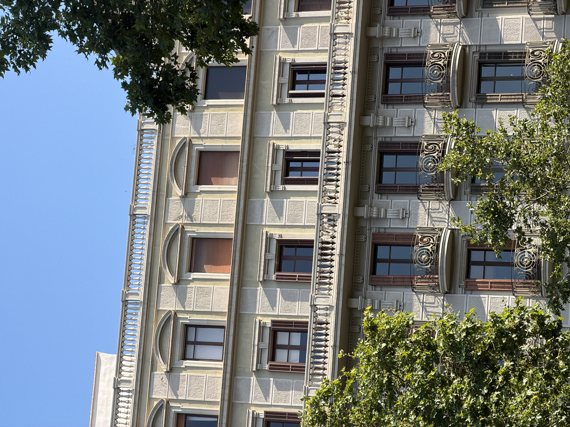 This is a close-up, low-angle shot of an ornate apartment building in Barcelona, Spain, bathed in bright, clear daylight, suggesting it is midday. The building's facade is a pale cream color with intricate decorative elements. Vertical bands of relief carvings frame rows of windows, each with dark wooden shutters. Some windows have decorative, wrought-iron balconies with circular, ornate designs. Trees with lush green leaves are visible in the foreground and upper left of the frame, partially obscuring the building and adding a natural element to the urban scene. The sky is a vibrant, cloudless blue, indicating fair weather. The image captures a sense of architectural detail and the interplay of light and shadow on the building's textured surface. No people or text are visible in the image.