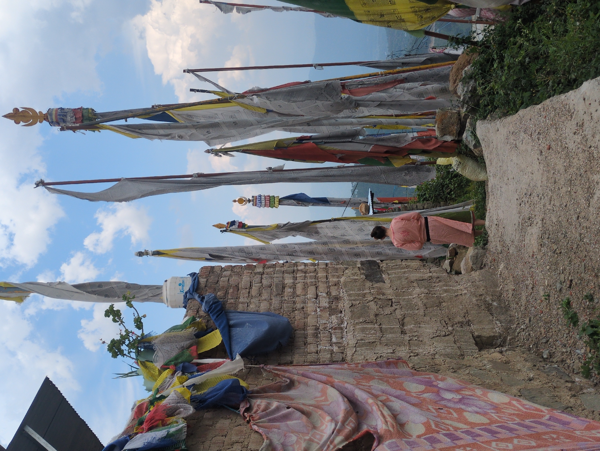 This image depicts an outdoor scene with a person walking on a gravel path. The path is lined with numerous prayer flags, many of which are tattered and worn, strung between poles. The flags are predominantly white, but also include yellow, blue, and red. In the background, mountains are visible under a cloudy sky. The environment suggests a high-altitude, possibly mountainous, location. A brick wall with a decorative cloth hanging on it is present to the left of the path. The person is wearing a light pink or peach-colored garment. The scene appears to be in a place of religious or cultural significance, given the abundance of prayer flags. The overall impression is one of a spiritual or contemplative atmosphere. The provided information states the location is the city of Doh, India, which is likely a misunderstanding, as there is no city named Doh in India. It is possible this refers to a location within the Indian Himalayas or a region associated with Buddhist culture.