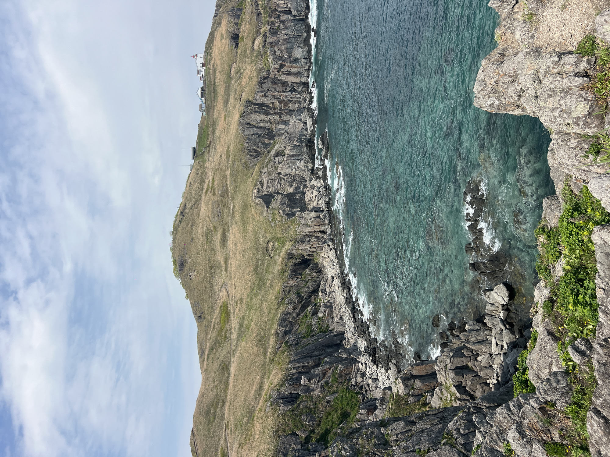 The image presents a view of a coastal cliff landscape. The foreground features a rocky outcrop with some green vegetation. The water along the shore is turquoise and relatively clear, with waves visible. A hillside covered in grass and some exposed rock faces rises from the shoreline, sloping upwards towards the top left of the frame. Buildings can be seen at the top of the hillside. The sky is overcast. The scene is of Mayak Byusse, Russia.