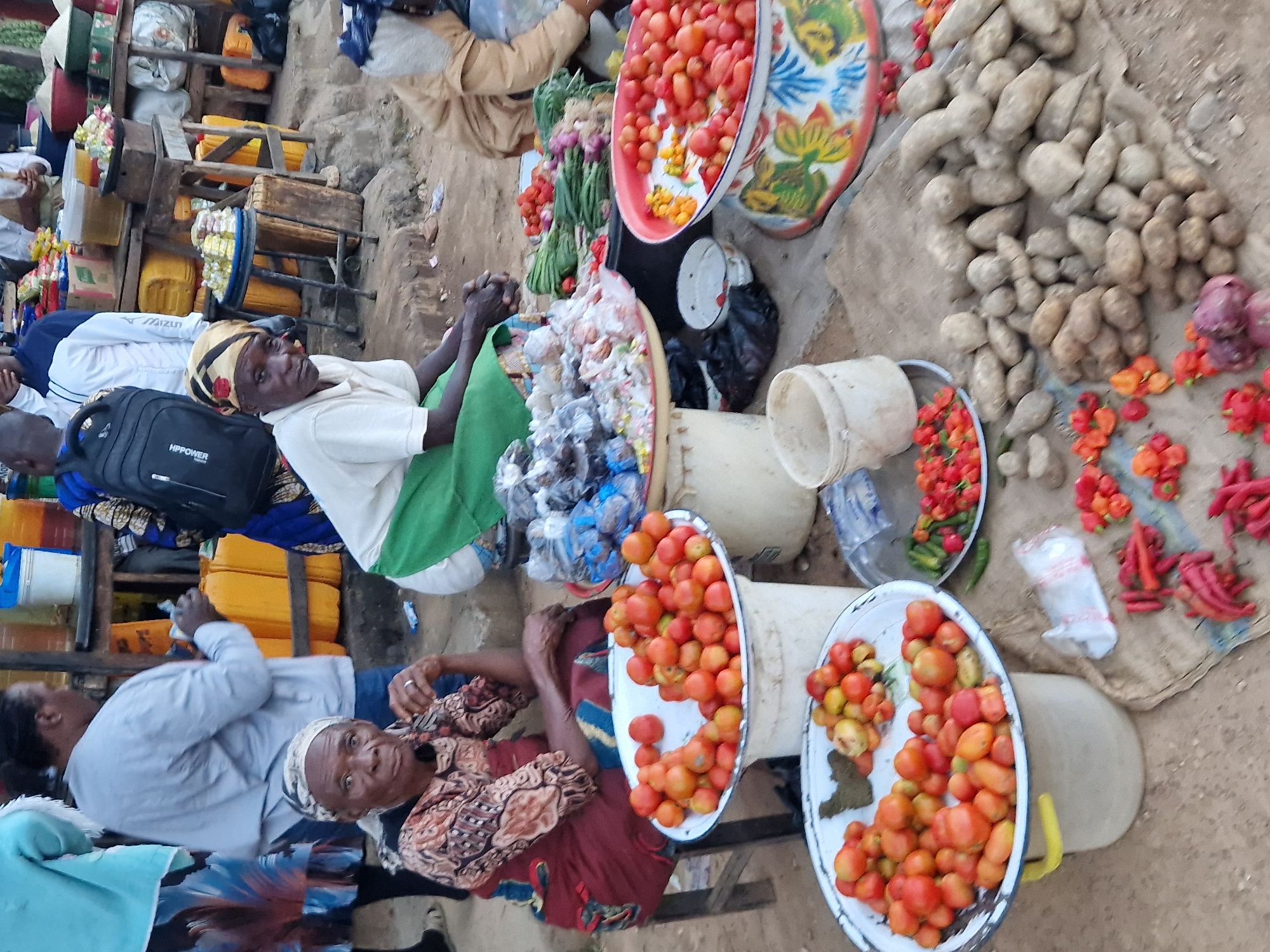 In Kabong, Nigeria, a busy market scene unfolds as individuals engage in commerce. A woman, likely a vendor, sits on a stool wearing a patterned top and a maroon skirt, her hand resting on her chin. In front of her are large white basins filled with ripe red tomatoes. Another woman, seated in the foreground, also wears a patterned headscarf and a white t-shirt, her hands clasped. A collection of yams and other root vegetables are piled on a burlap sack, while small red chilies are scattered across the ground. In the background, yellow jerrycans suggest liquid goods are also for sale. The market appears to be outdoors, with a dirt and gravel ground. People in the background are seen interacting with stalls, indicating a lively marketplace.