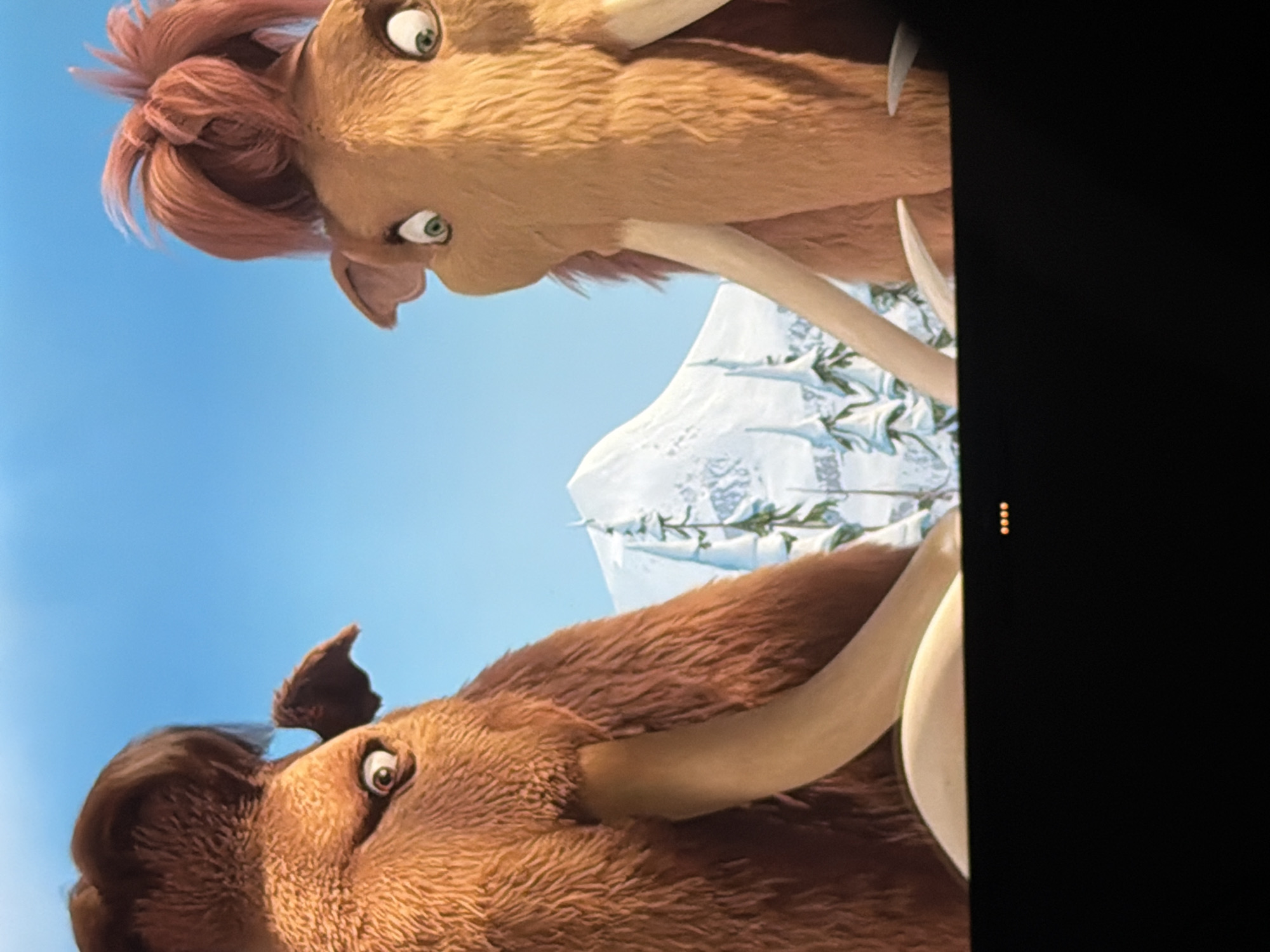 Two animated mammoths are in the foreground. The mammoth on the left has dark brown fur and a dark brown hairstyle. The mammoth on the right has orange-brown fur and a pinkish-orange hairstyle. Both mammoths have large tusks. In the background, there is a snow-covered mountain and pine trees. The sky is blue. The image is captured from a low angle, looking up at the mammoths. The bottom of the image is black. There are four yellow dots in the bottom center of the image. The location is the city of Chaikovsky, Russia.